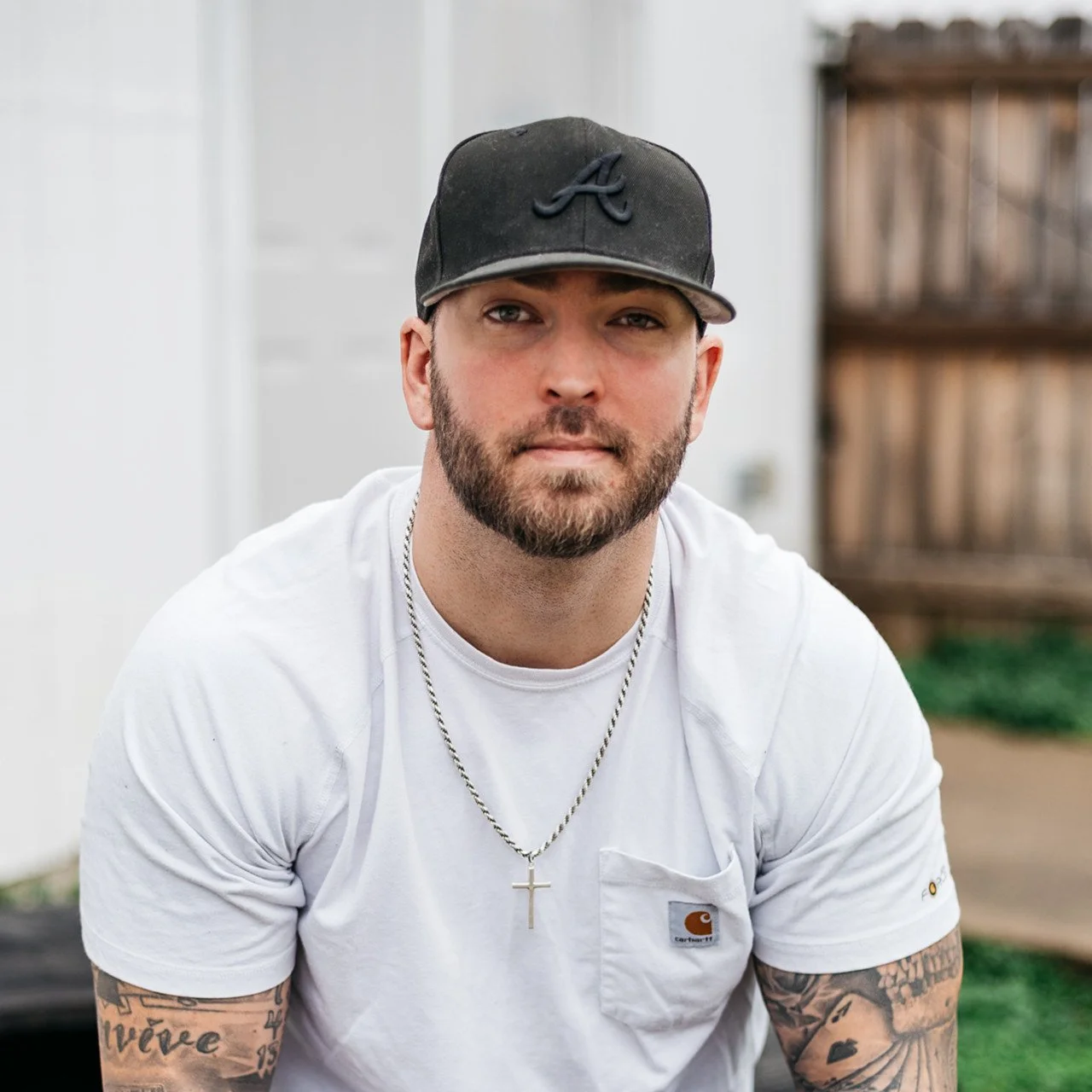 A young man with a beard wearing a black cap, white T-shirt, and a cross necklace, sitting outdoors in a backyard with wooden fence and white building in the background.