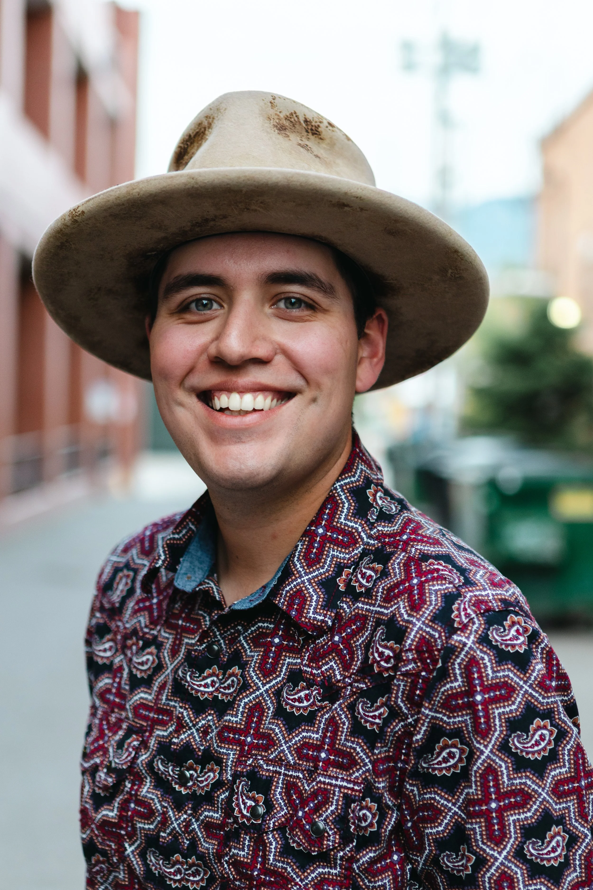 A man with a beard wearing a wide-brimmed hat, denim jacket, and plaid shirt stands in a field of tall grass with mountains in the background.