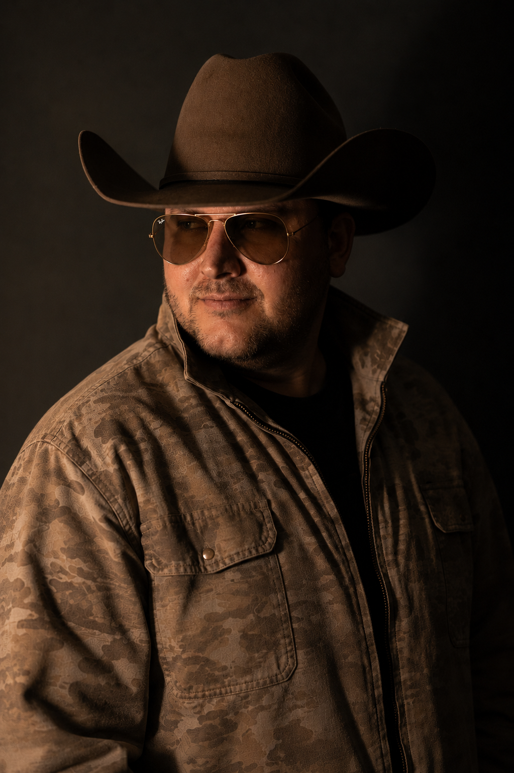 A man wearing a cowboy hat, tinted sunglasses, a camouflage jacket, and a black shirt against a dark background.