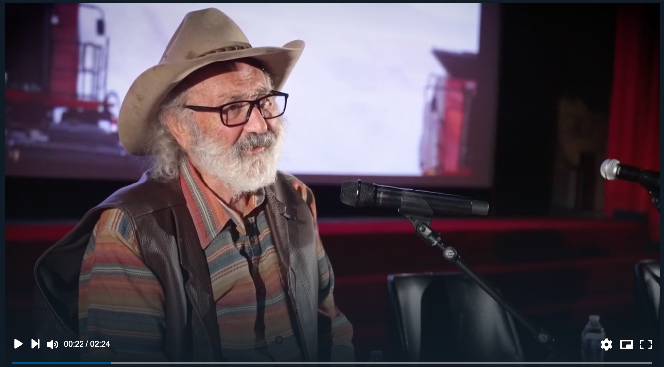 An elderly man with white hair and beard wearing a cowboy hat, glasses, a striped shirt, and a leather vest, sitting at a table with microphones in front of him, in a room with a large screen displaying an image of a red building.