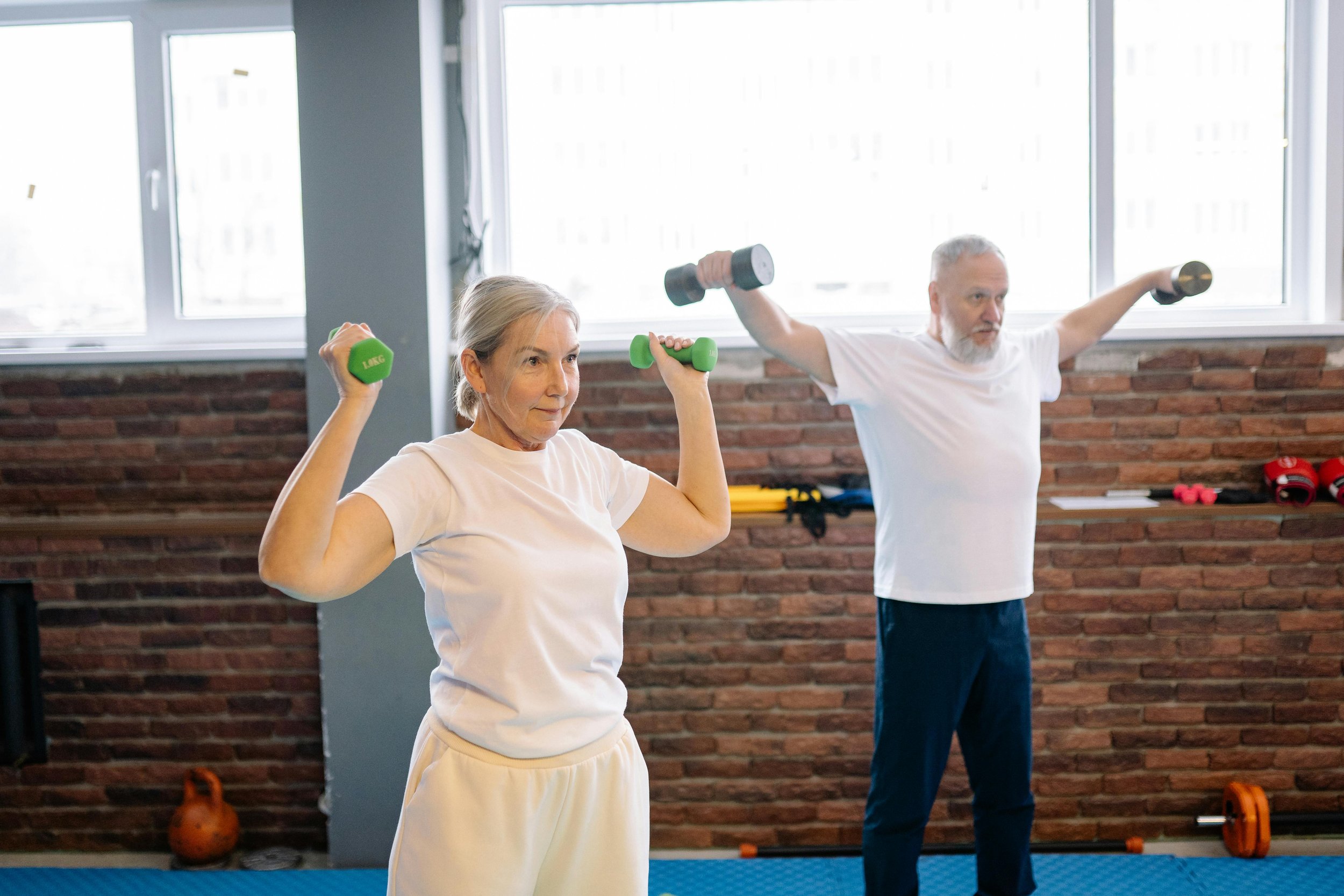 Older woman and man exercising with dumbbells in a gym with large windows and brick walls.