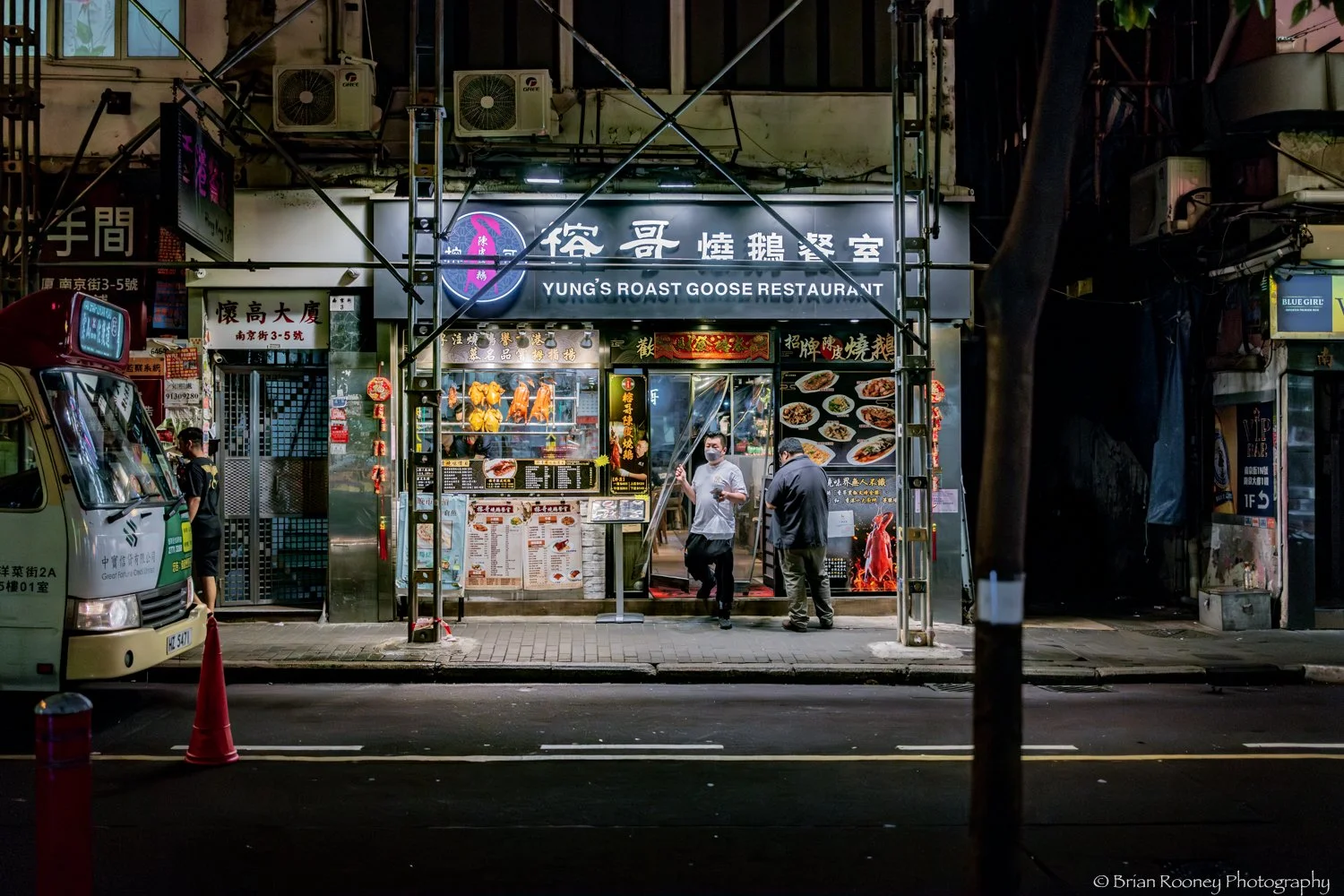 Nighttime street scene in front of Yung's Roast Goose Restaurant, with outdoor menu displays and people standing outside, illuminated neon sign in English and Chinese, parked vehicles on the street.