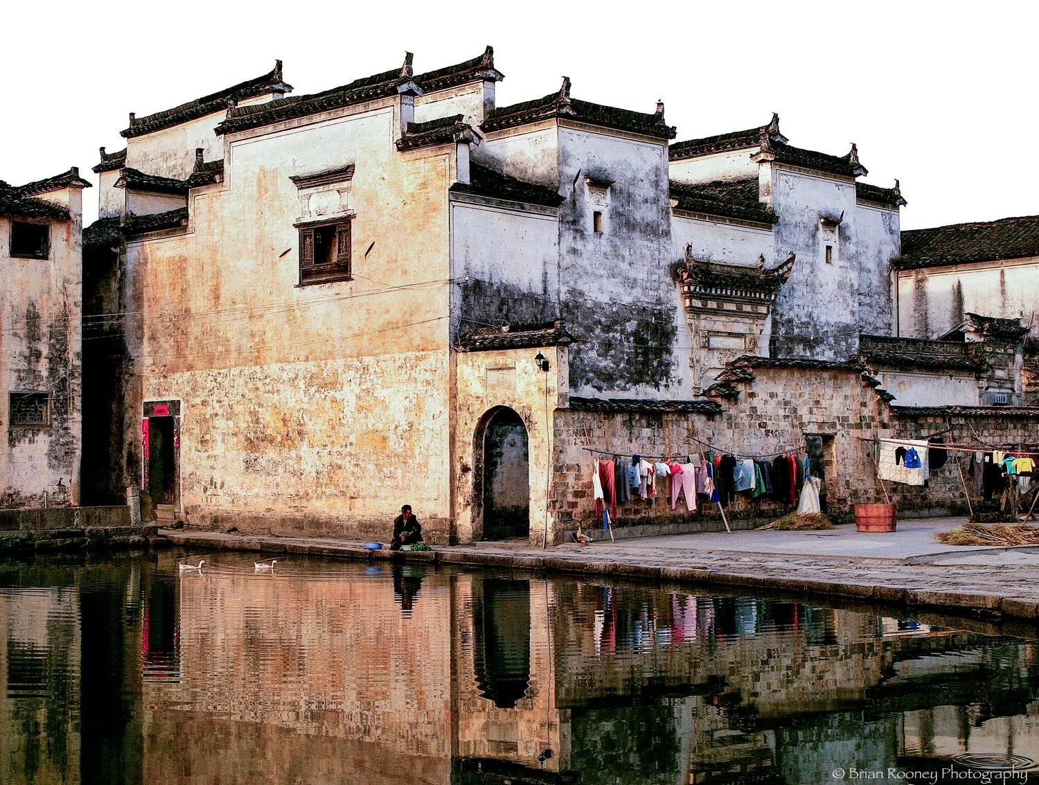 An old Chinese building with weathered walls, adjacent to a water body with ducks. Clothes are hanging on lines to dry along a stone wall, and a person is sitting near the water.