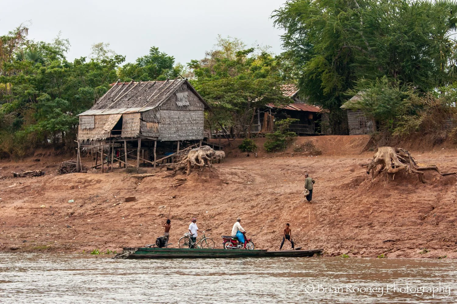 People on a boat near the riverbank of a rural area with wooden houses on stilts, trees, and large tree roots visible along the shoreline.