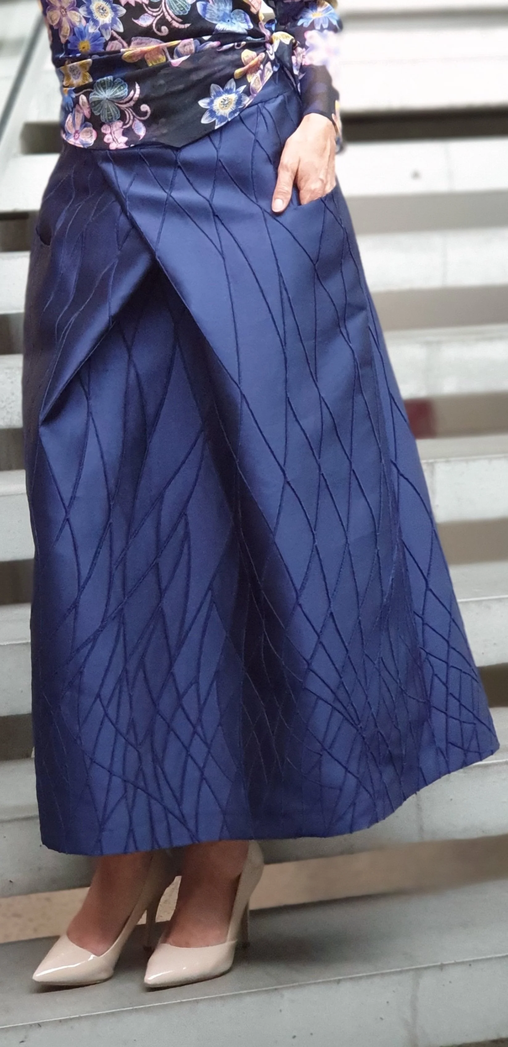 Frau trägt eine blaue, gemusterte Rockhose, helle High Heels, und ein schwarzes Oberteil mit Blumenmuster.