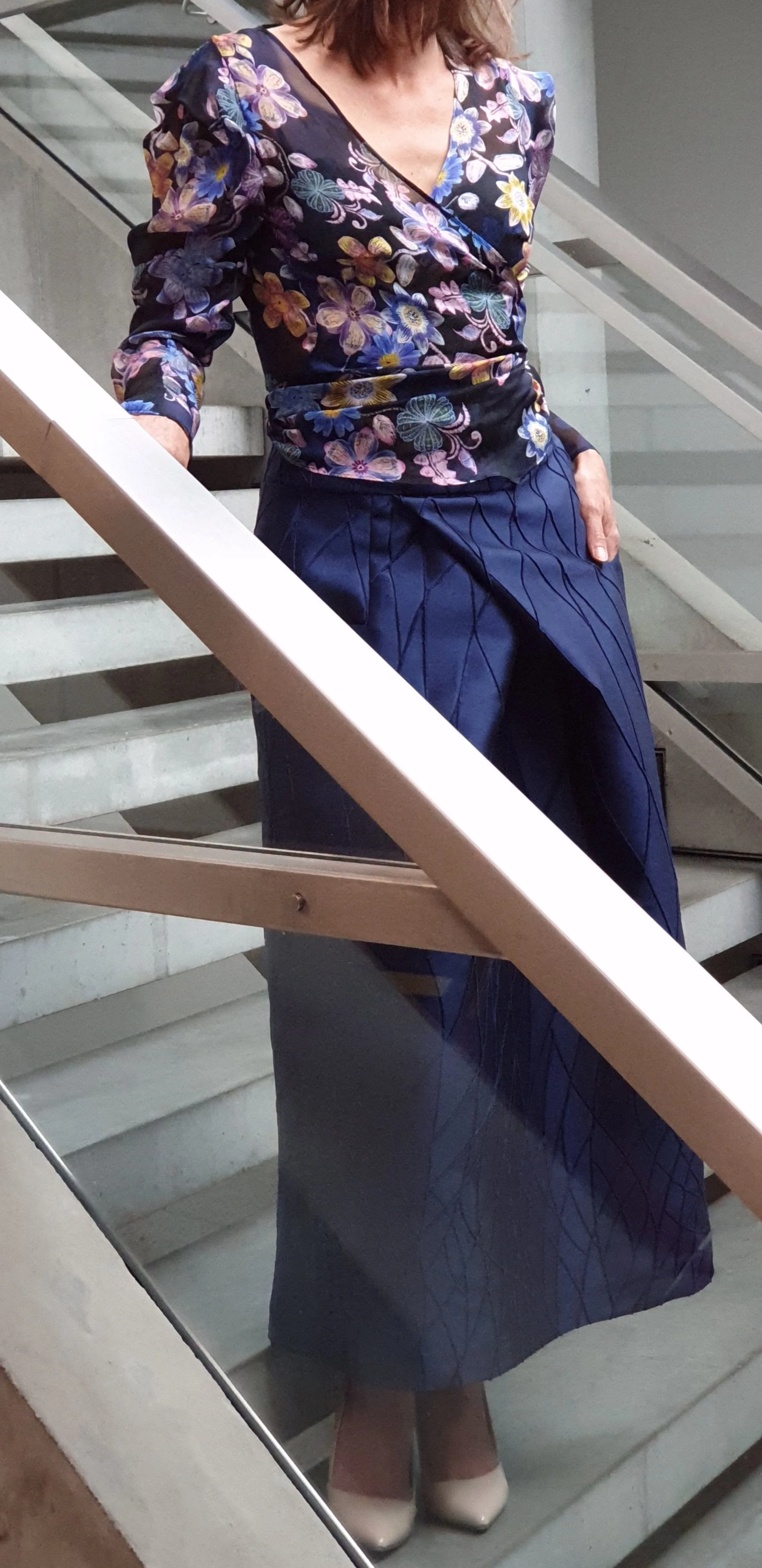 Frau in bunter Blumenbluse und blauer Plisseerock, stehend auf einer Treppe, mit Hand in der Tasche.