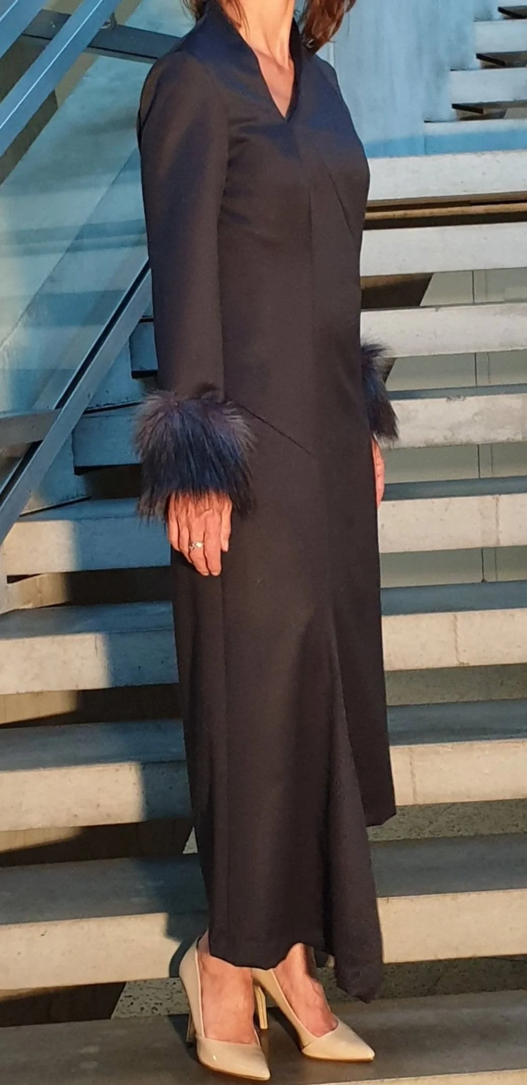 Frau in langem schwarzen Kleid mit Pelzbesätzen an den Ärmeln, weißen High Heels, stehend auf einer Treppe.