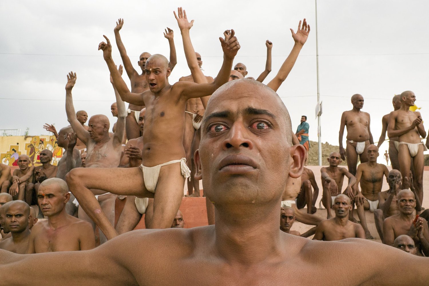A mass initiation rite: dozens of shaven-headed men in white loincloths with arms flung upward, and in the immediate foreground one man's face fills the frame, eyes bloodshot and streaming, a tear on his cheek