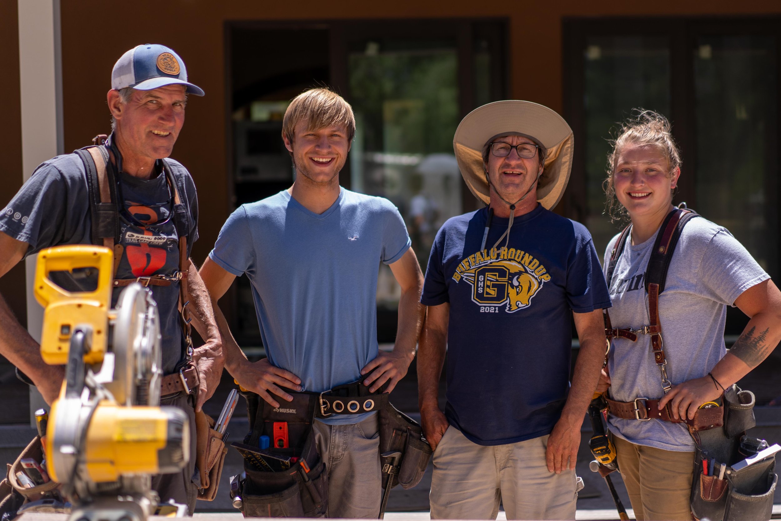 Group of four construction workers smiling outdoors, wearing casual clothing and tool belts, standing in front of a building with a saw in the foreground.