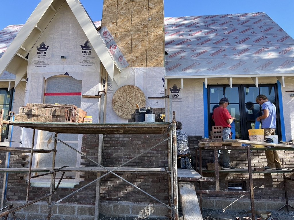 Construction site with workers building a brick facade on a house. Scaffolding in front and roofing materials visible.