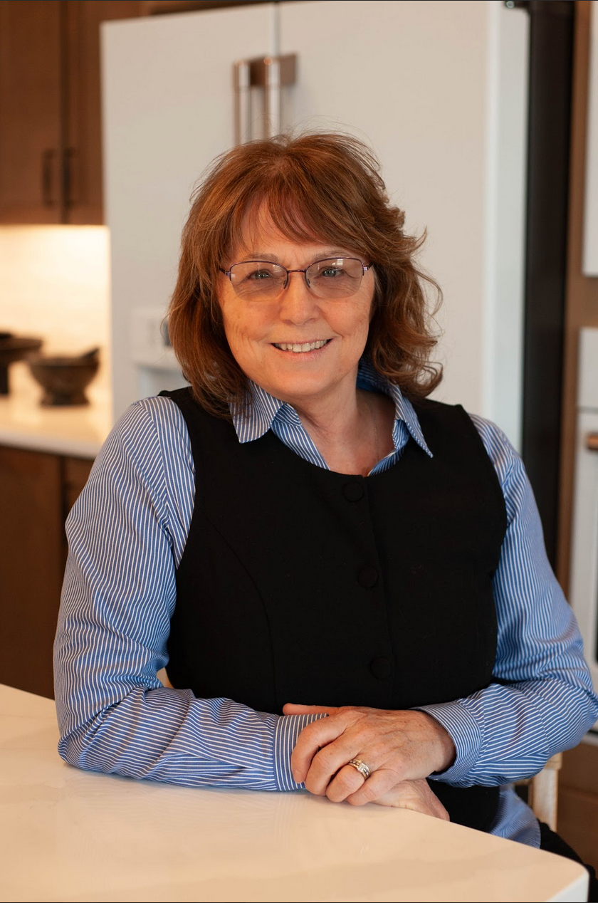 Smiling woman with light brown hair, wearing a blue shirt, standing in a kitchen.