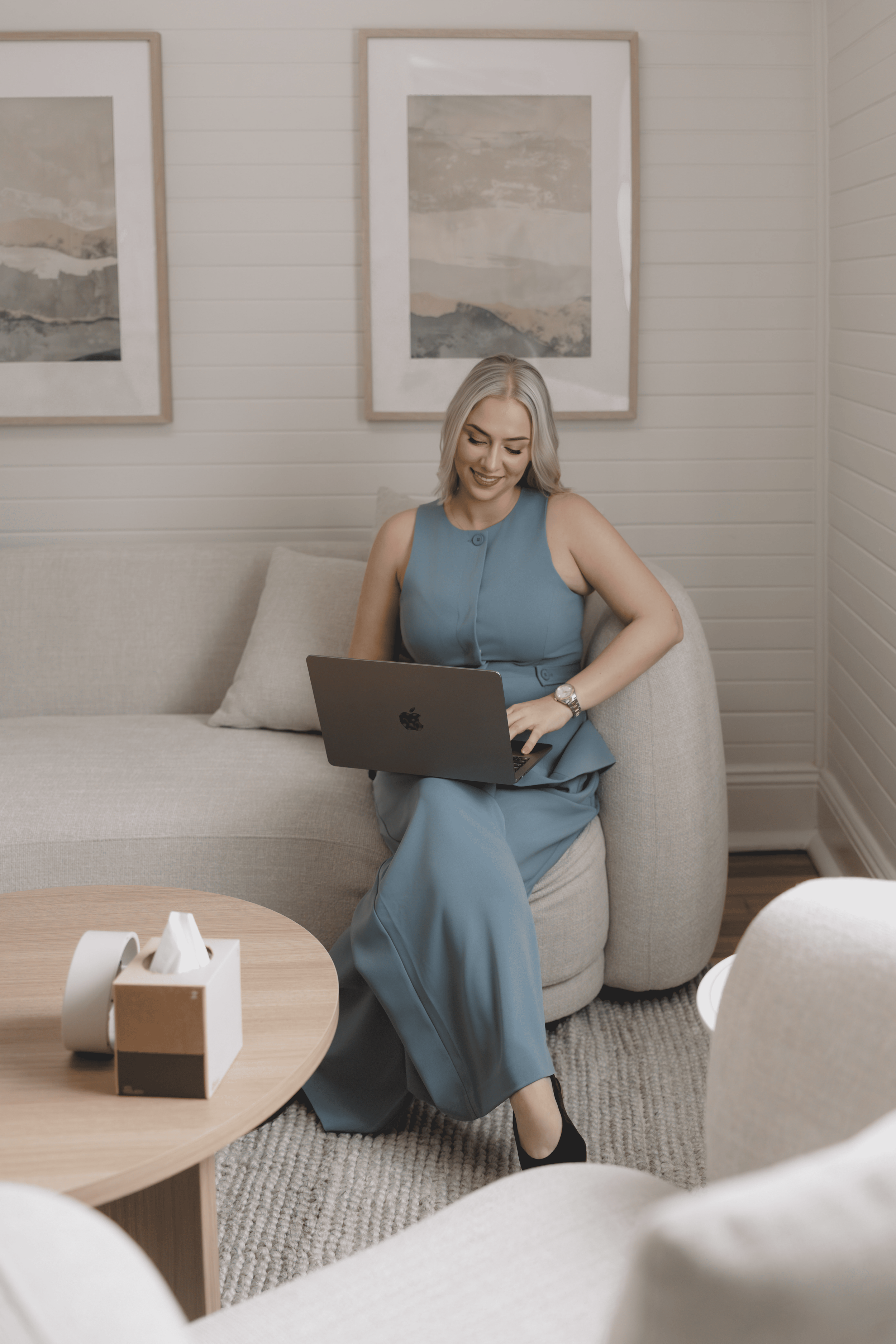 A psychologist, dressed in a blue sleeveless dress, sitting on a beige sofa in a modern living room, working on a laptop, with a smile.
