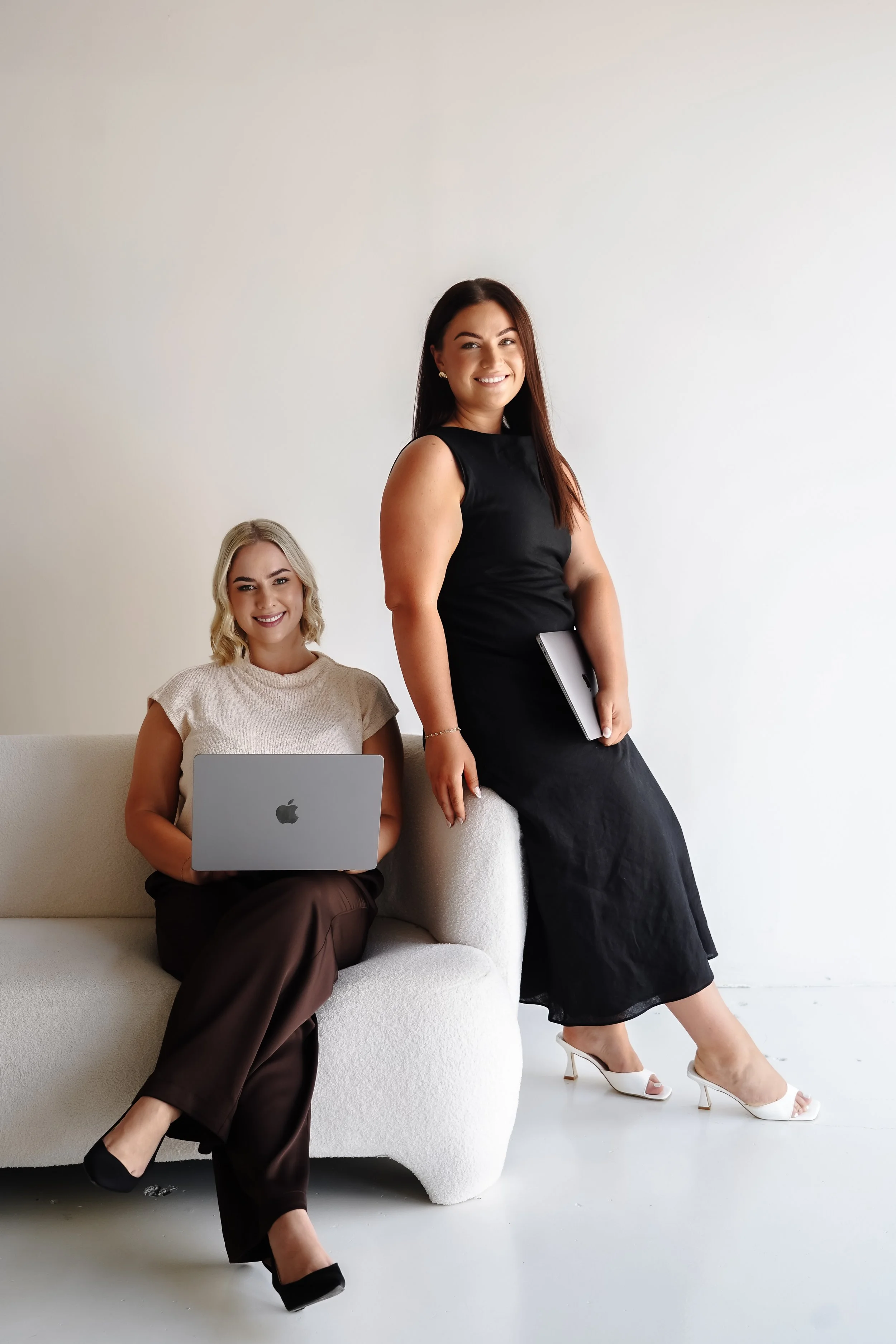 Two therapists with laptops and a tablet in a minimalistic room with a white wall. One woman is sitting on a white couch, holding a silver laptop, wearing a beige top and brown pants. The other woman is standing, holding a tablet or notebook, wearing