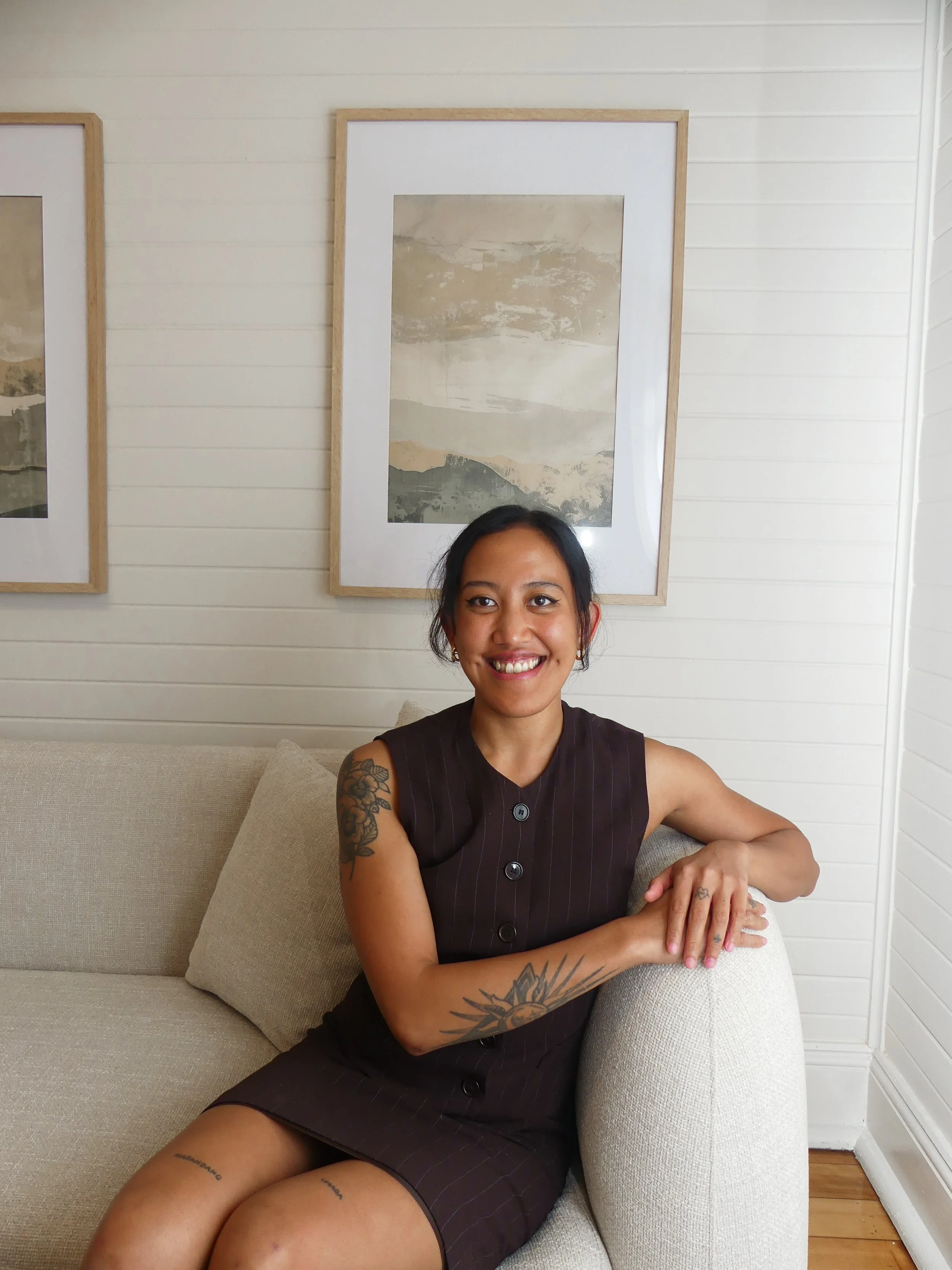 A young woman with tattoos on her arm and legs, sitting on a beige sofa in a room with white wood-paneled walls and framed artwork behind her. She is smiling, wearing a sleeveless black pinstripe dress.