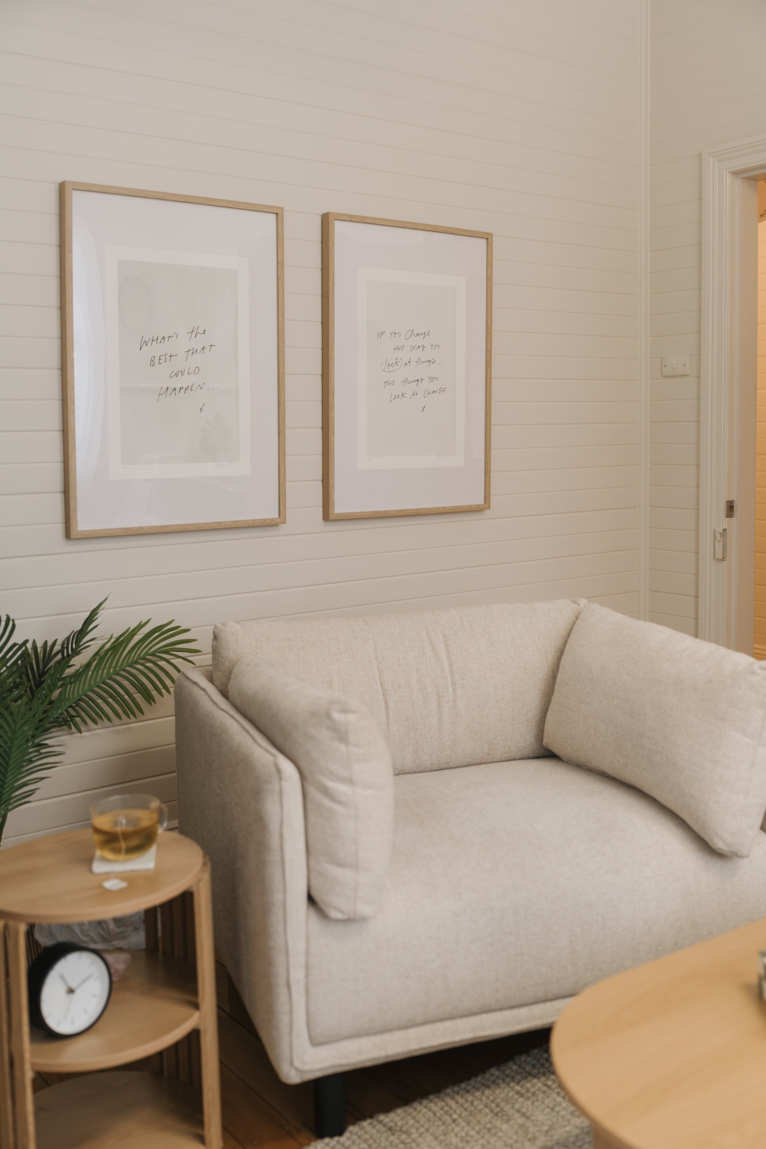 Living room with a beige couch, plant in the corner, a clock on the side table, and two framed quotes on the white, wooden wall.