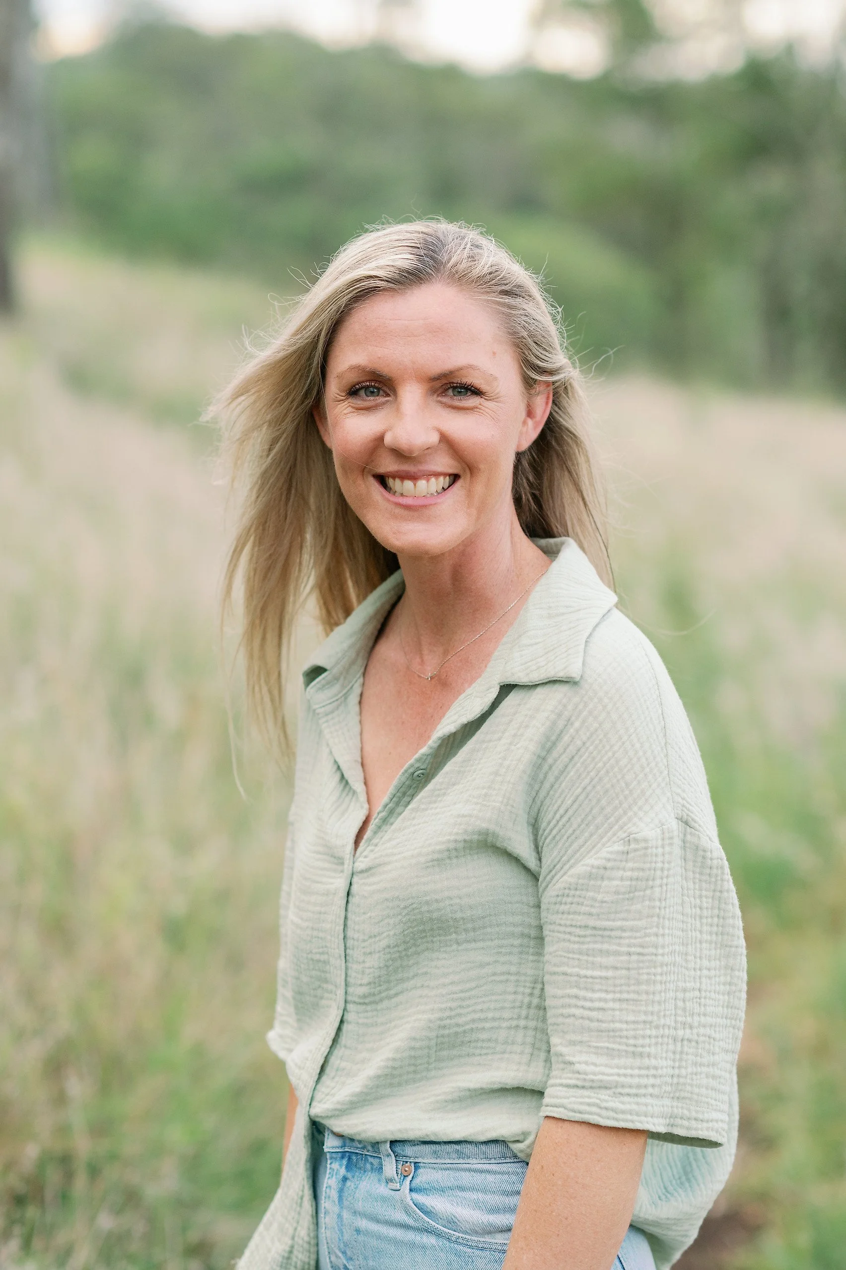 A woman with blonde hair smiling outdoors, wearing a light green shirt and blue jeans, with a blurred green natural background.