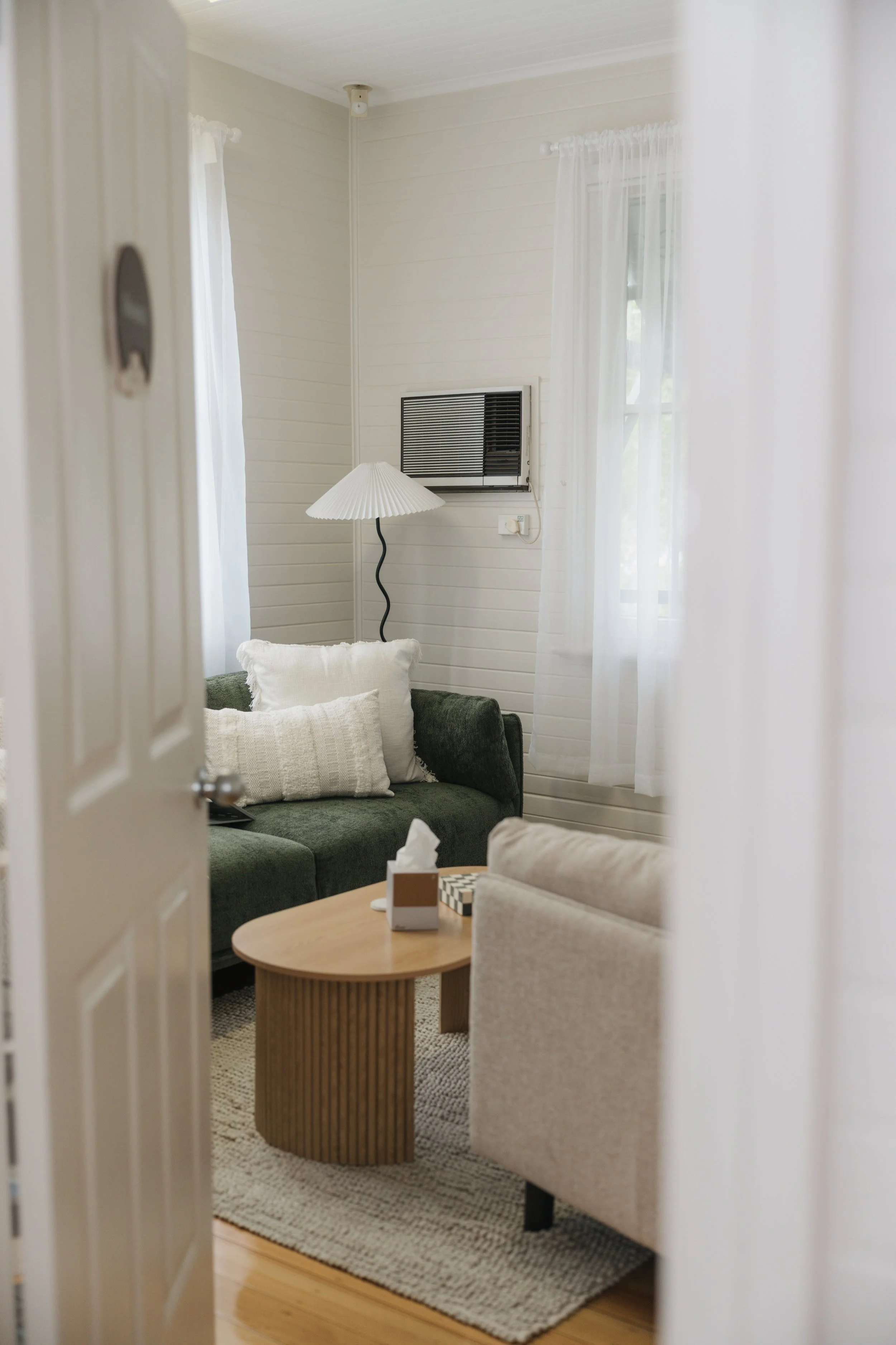 Living room with white paneled walls, white curtains, a green sofa with white pillows, a round wooden coffee table with a tissue box and a black and white checkered item, a beige armchair, a floor lamp with a pleated shade, and an air conditioning unit on the wall