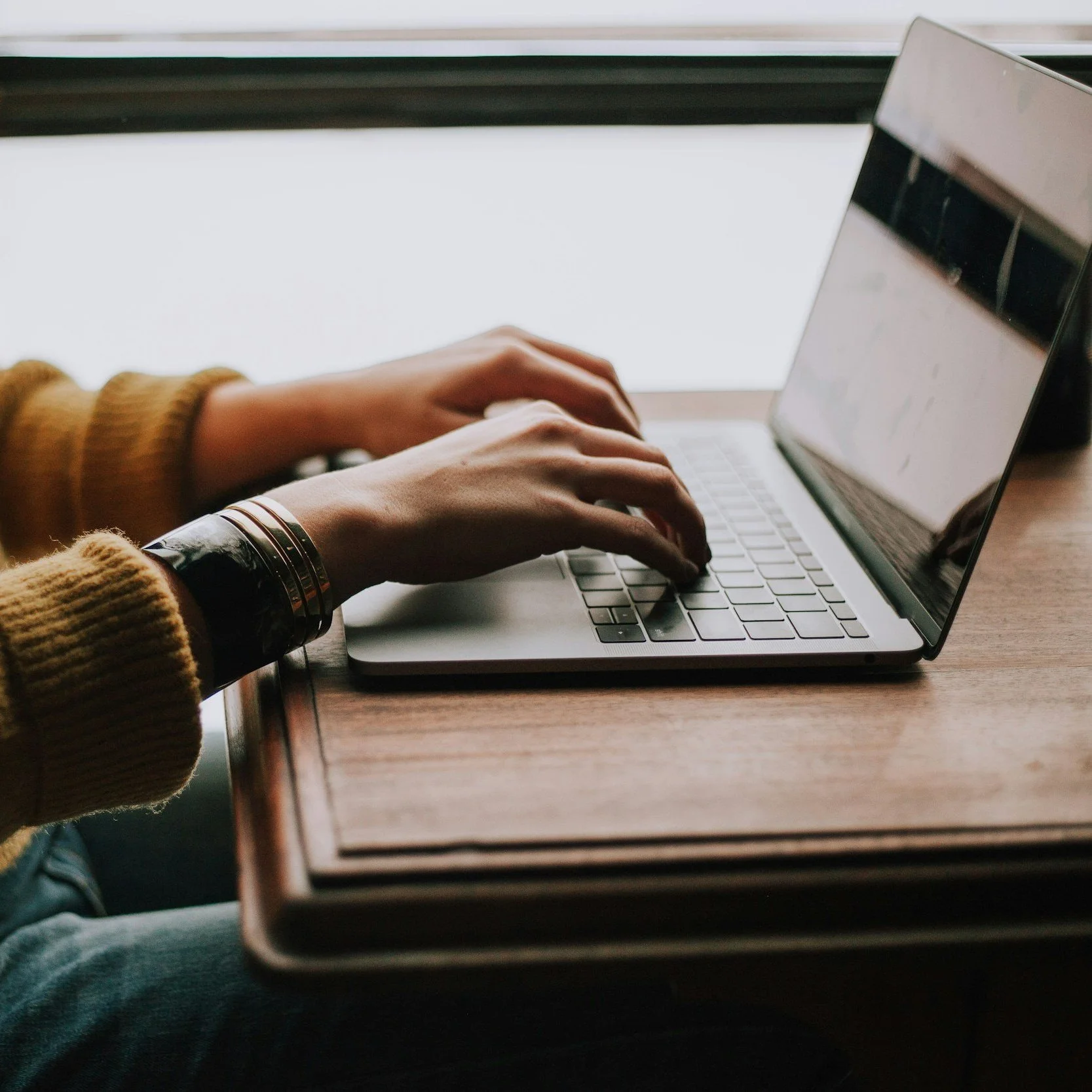 Person using a laptop at a wooden table near a window.