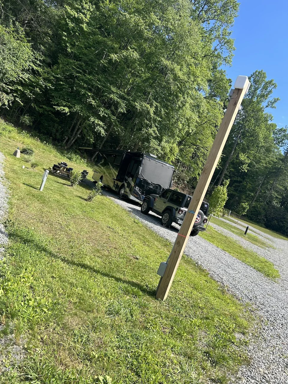 A grassy outdoor campsite with a large black RV and a smaller yellowish-green Jeep parked on gravel. There is a wooden utility pole in the foreground, some small plants and a picnic table nearby, and dense green trees under a clear blue sky.