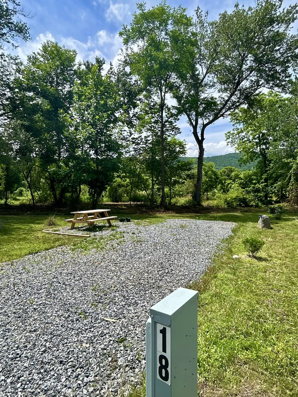 Gravel pathway next to a picnic table in a grassy area with trees and a blue sky.
