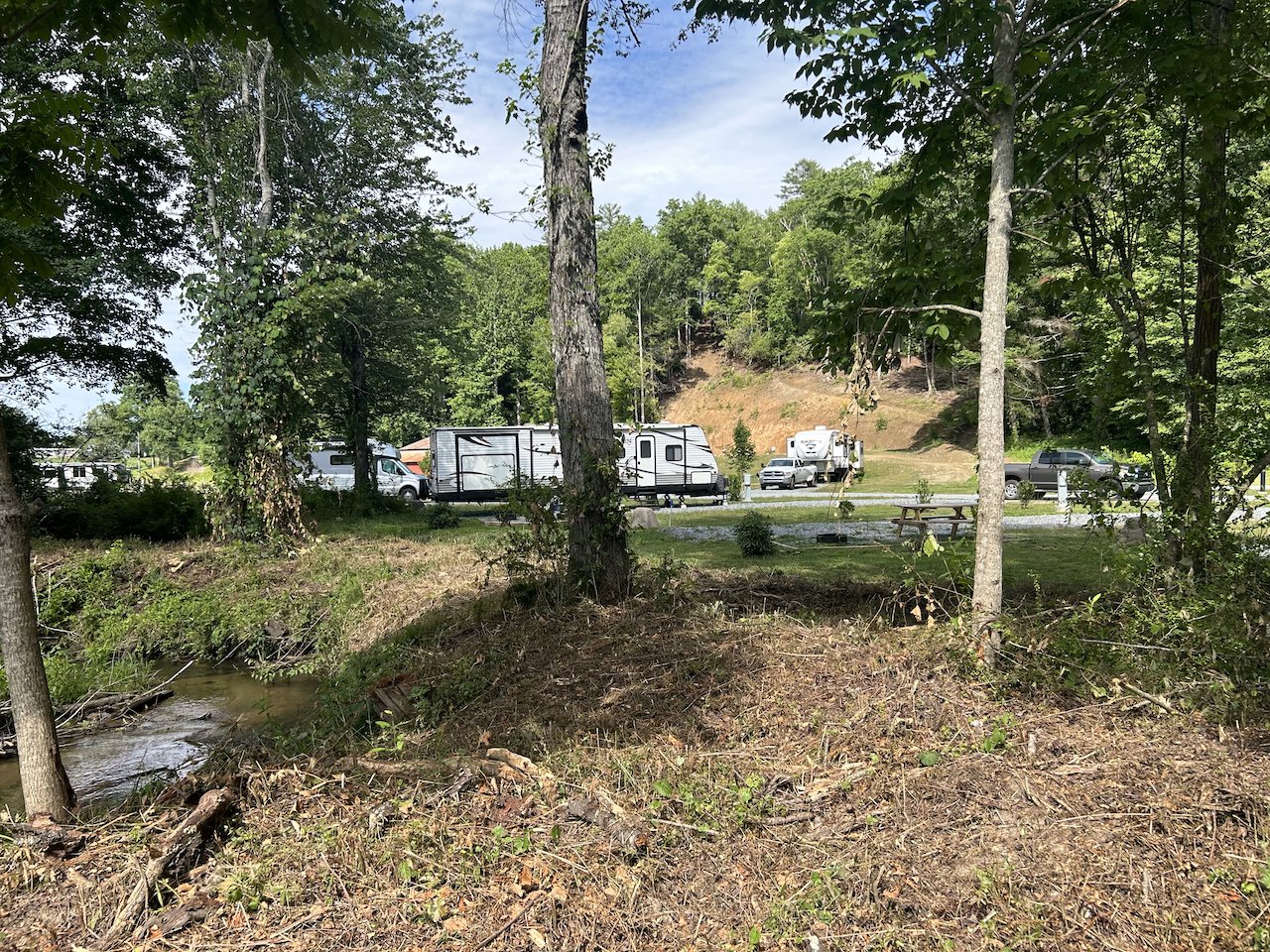 A wooded area with a small creek in the foreground and multiple RVs and trucks parked along a road in the background.