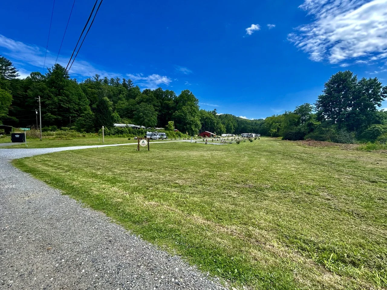 A grassy field with a gravel path on the left, lined with trees and power lines in a rural area under a bright blue sky with clouds.