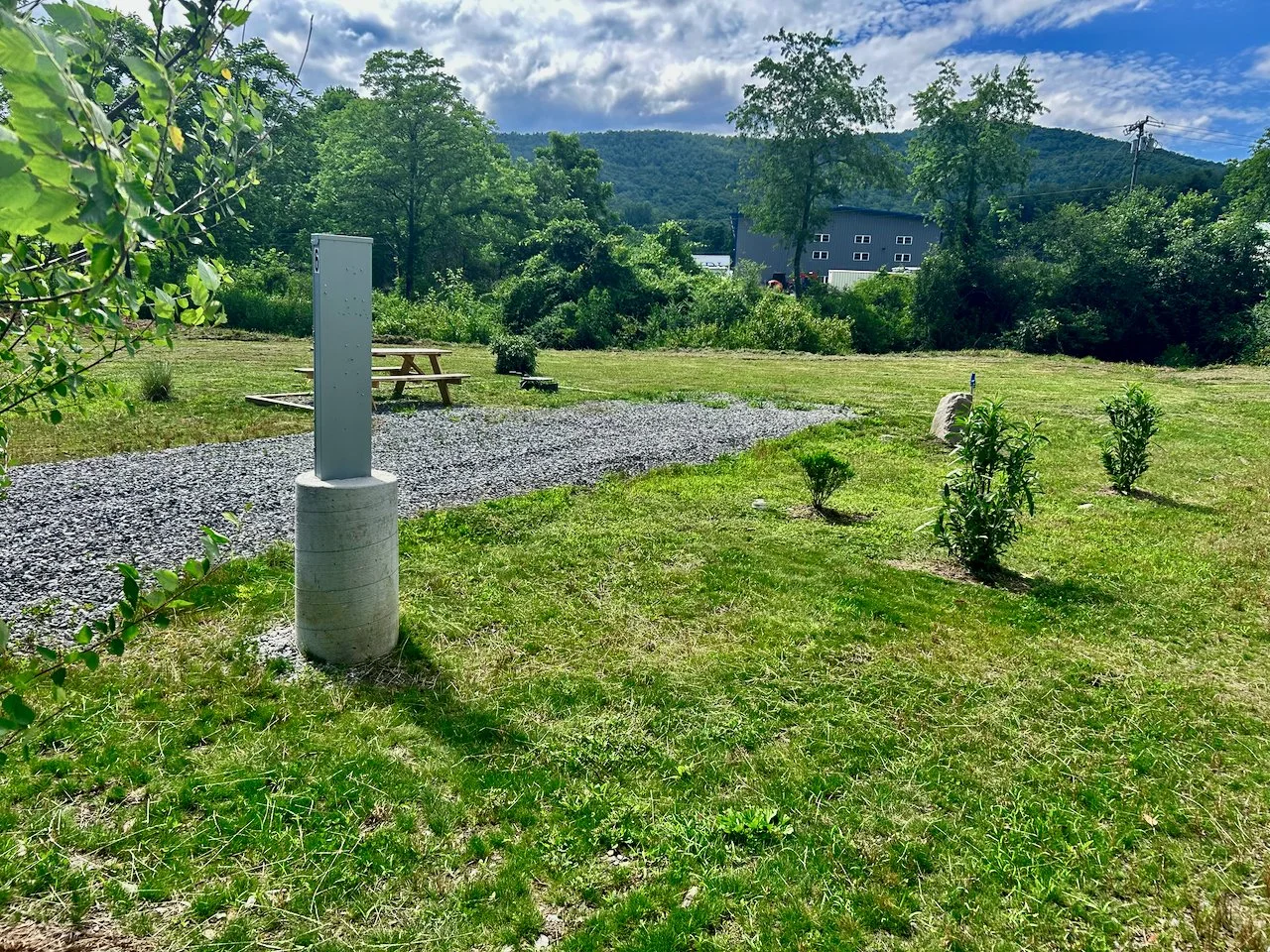 A grassy park area with small plants in the foreground, a gravel pathway, a picnic table, and a water fountain. In the background, there are trees, a building, and a mountain under a partly cloudy sky.
