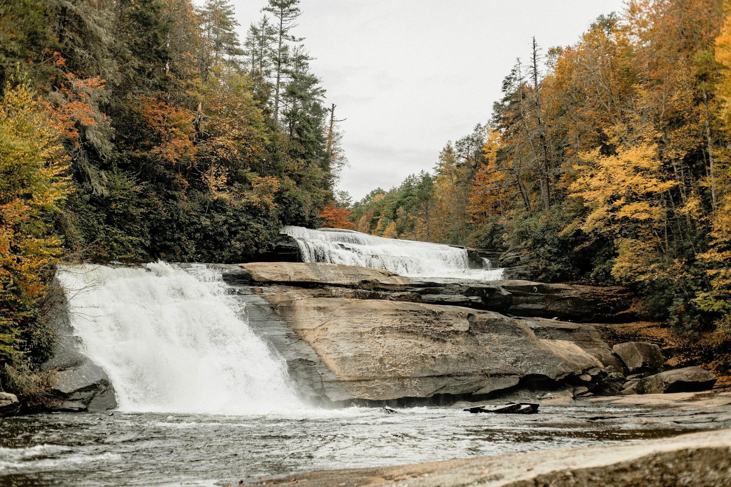 A multi-tiered waterfall flowing over rocks amidst a forest with autumn foliage.