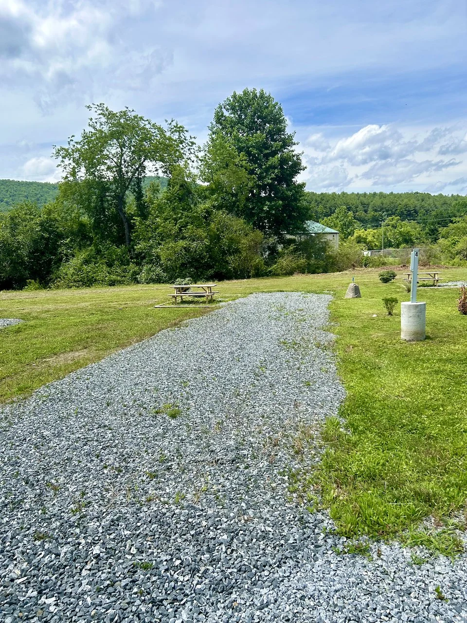 A gravel pathway winding through a grassy campground area with picnic tables, trees, and a blue sky with clouds.