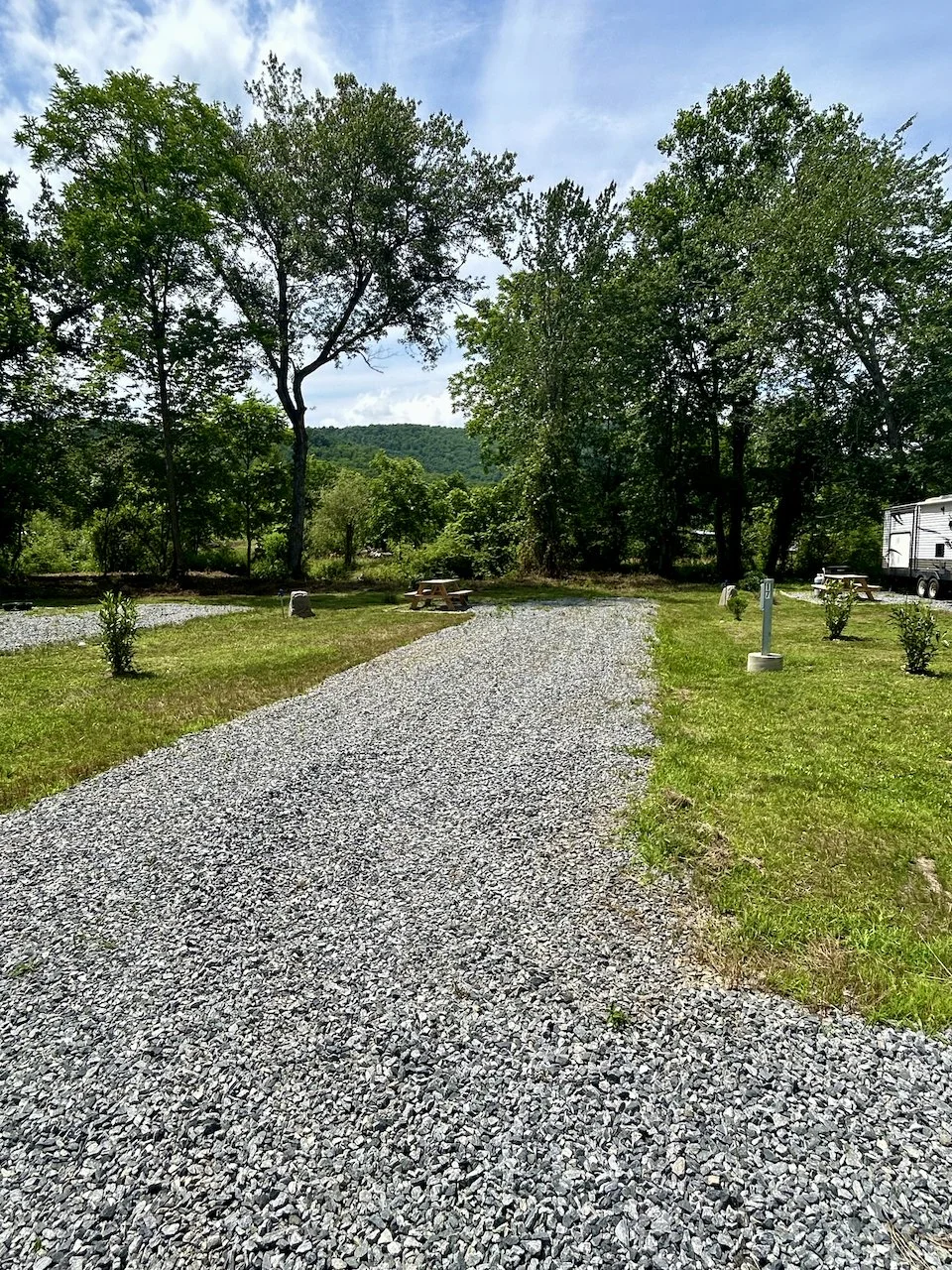 Gravel pathway in a grassy area with trees, picnic tables, and a red and white trailer in the background.