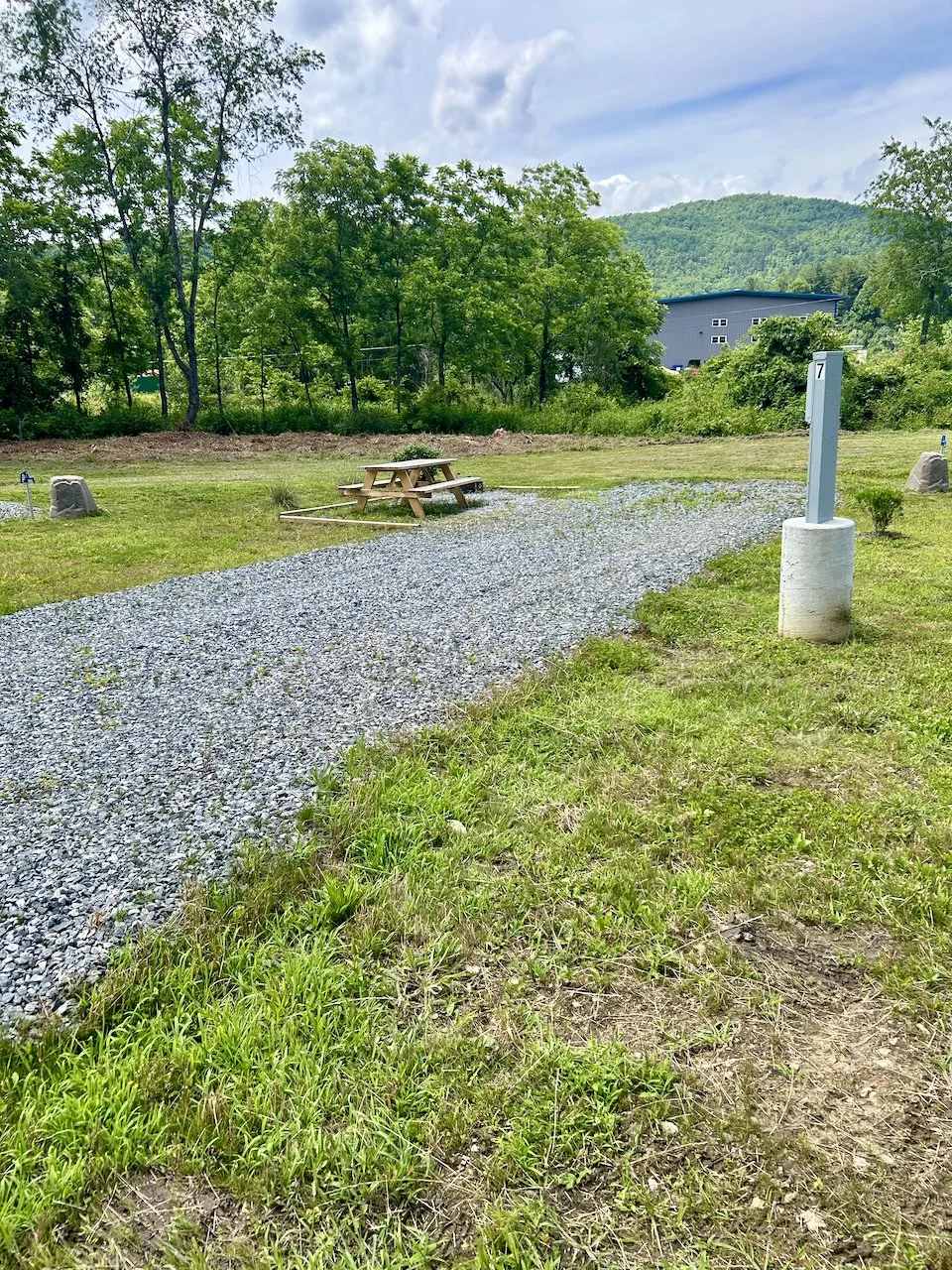 Gravel pathway with a wooden picnic table, trees, and a building in the background, and a grassy area with a marked post labeled '7' in a rural setting.