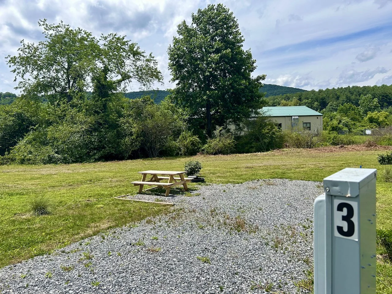 A rural outdoor scene with a gravel parking area, a picnic table, a small shrub, and a large green canopy of trees in the background. A building with a green roof is visible behind the trees, and a numbered utility post with the number 3 is in the fo