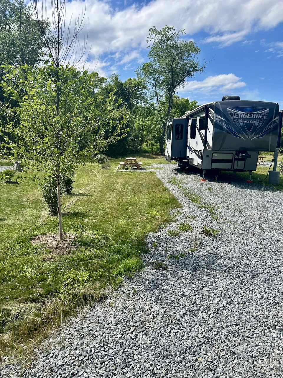 A mobile RV named 'Vengeance' parked on a gravel pad in a grassy area with trees and a picnic table, under a blue sky with some clouds.