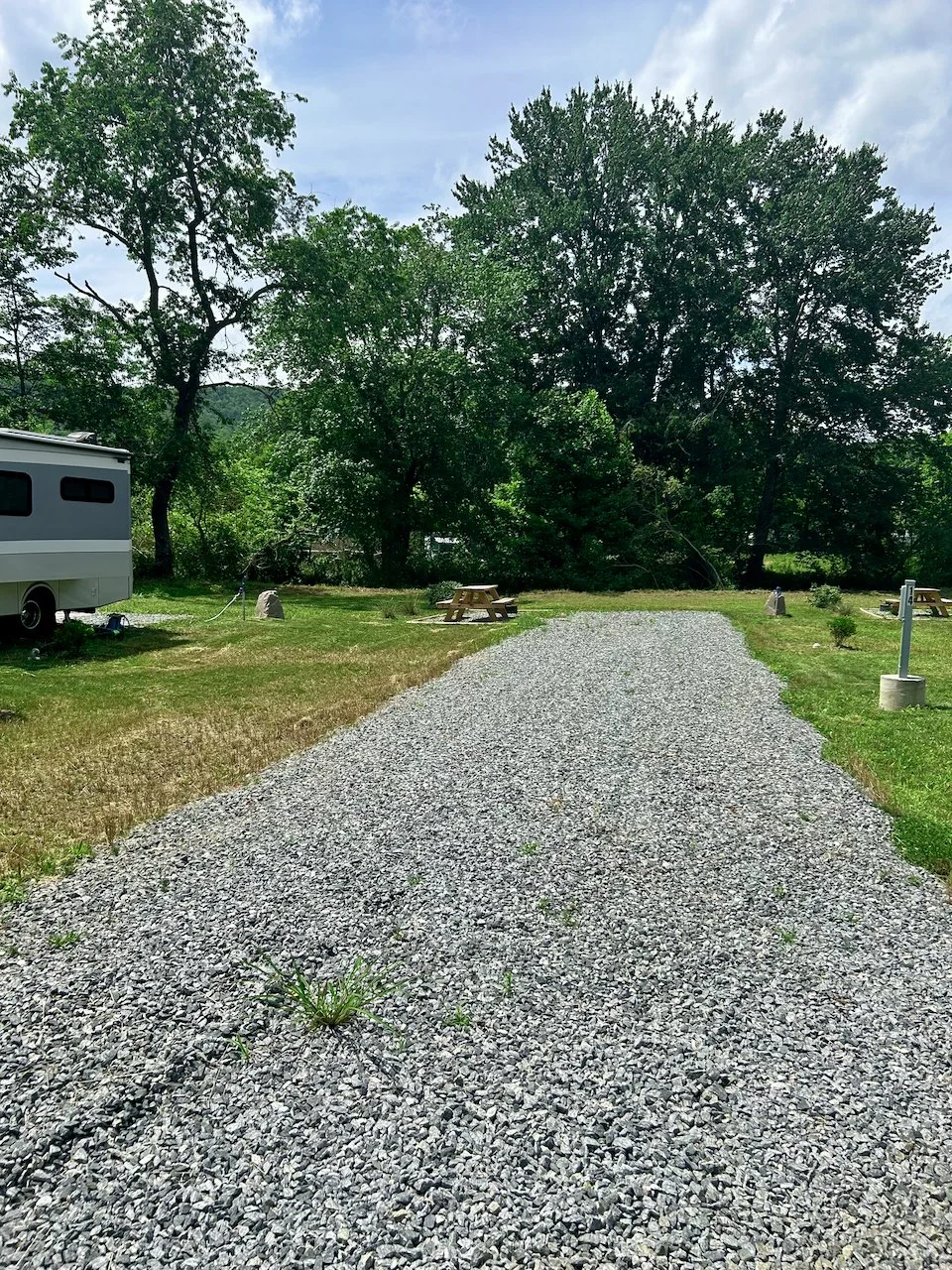 Gravel driveway leading to a wooded area with large trees, picnic tables, and a camper on the left side, under a partly cloudy sky.