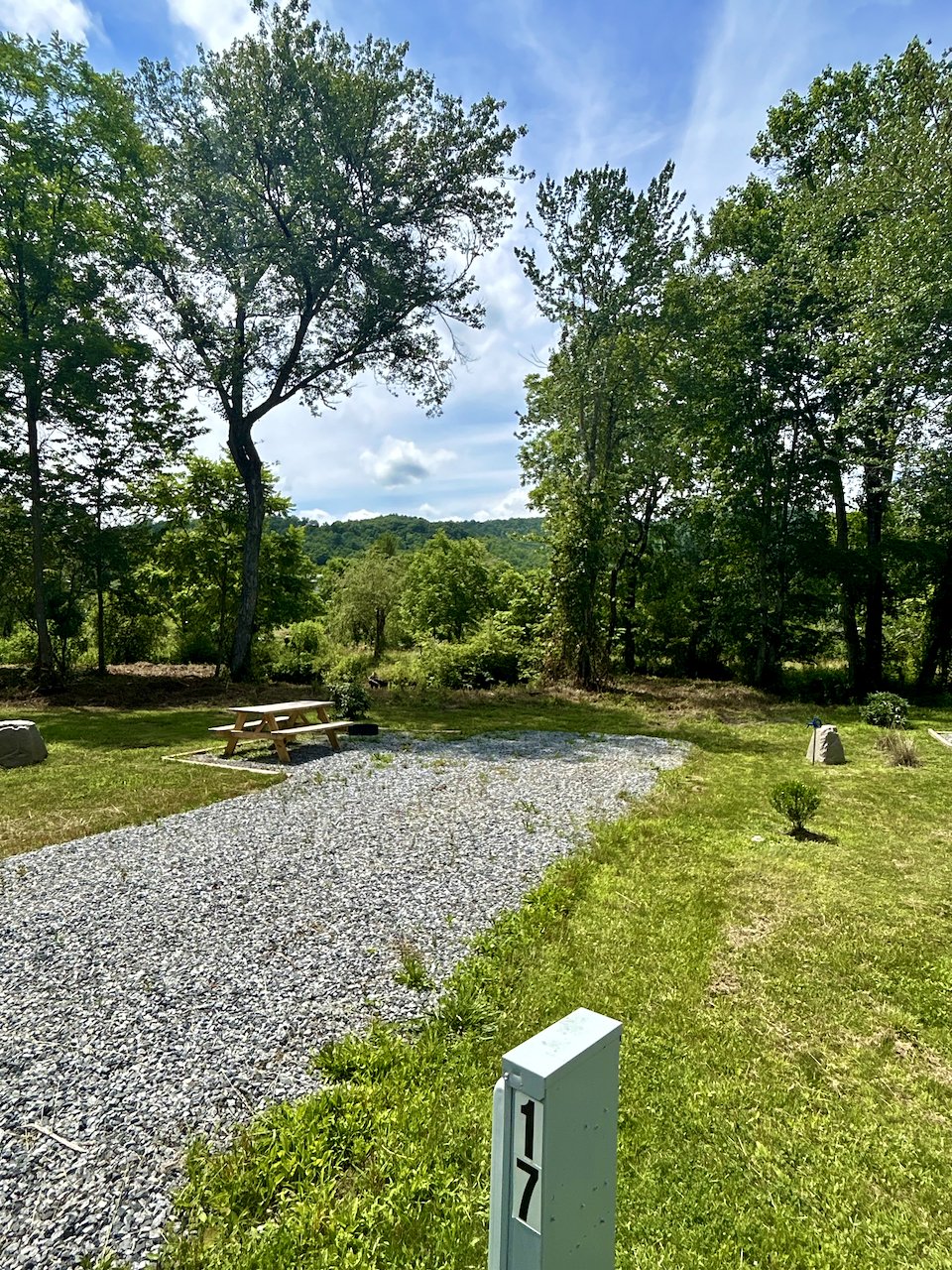 A scenic outdoor landscape with a gravel pathway, picnic table, lush green trees, and a clear blue sky with clouds in the background.