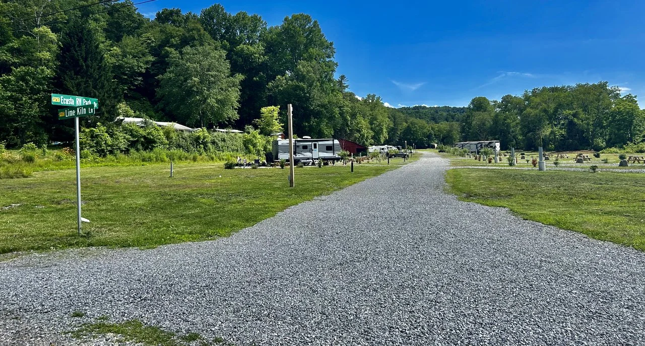A gravel road in a campground with tents and RVs on both sides, surrounded by trees and a partly cloudy blue sky.
