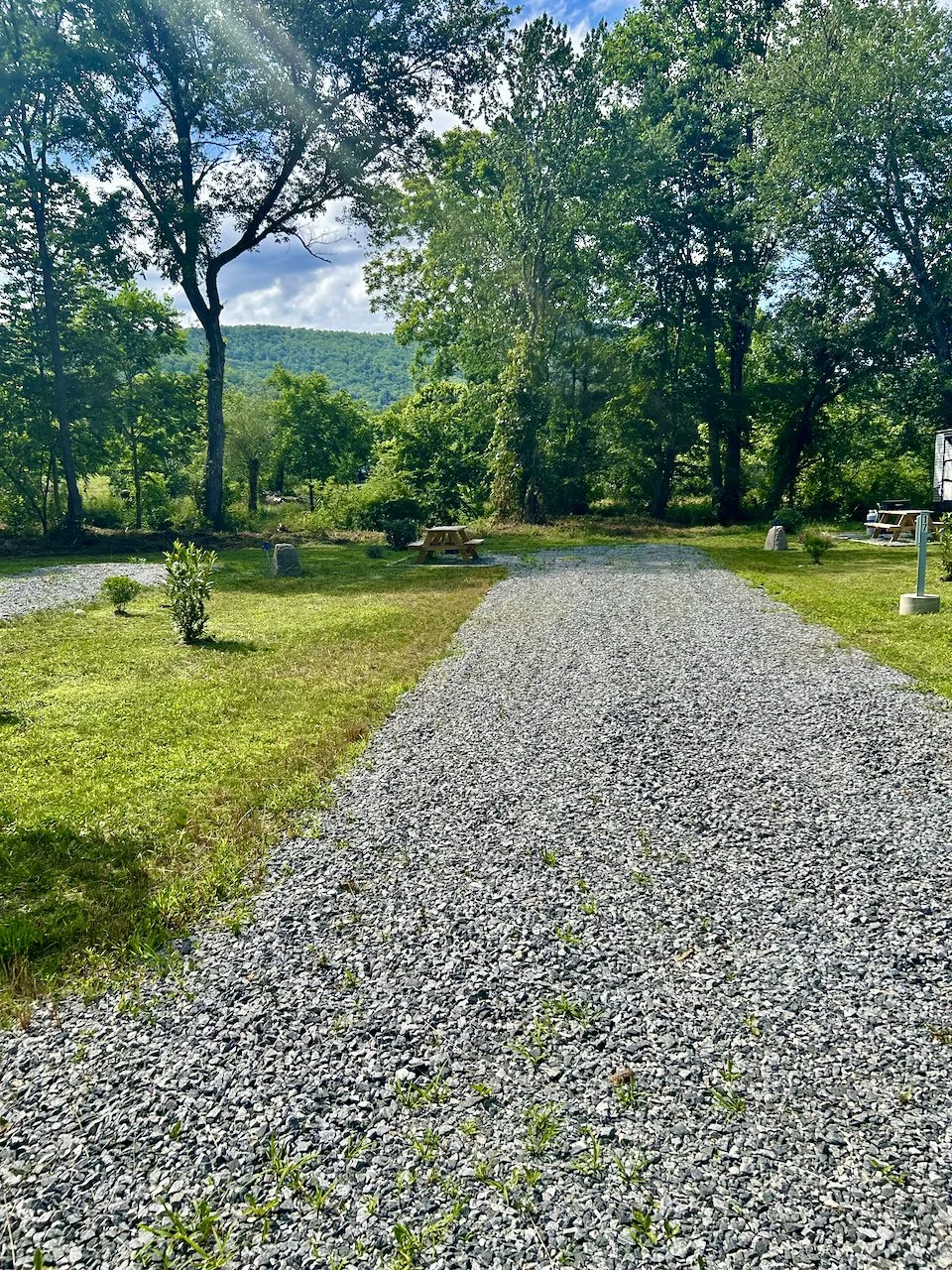 Gravel pathway in a grassy area surrounded by trees and bushes, with picnic tables and a water spigot visible in the background, under a partly cloudy sky.