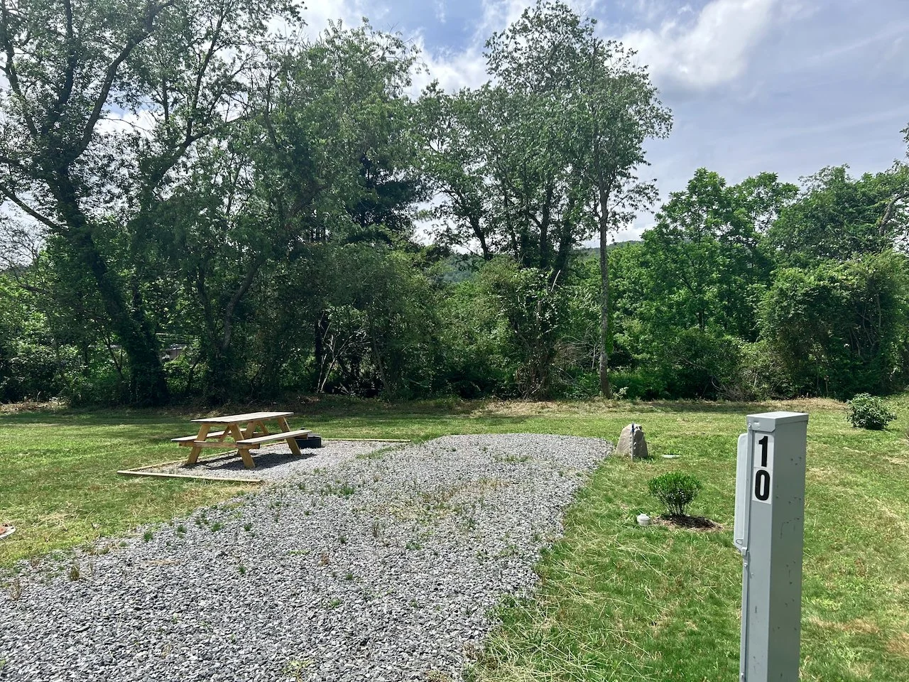 A gravel pathway leading to a wooden picnic table on a grassy area, with trees and bushes in the background, and a gray utility box with the number 10.