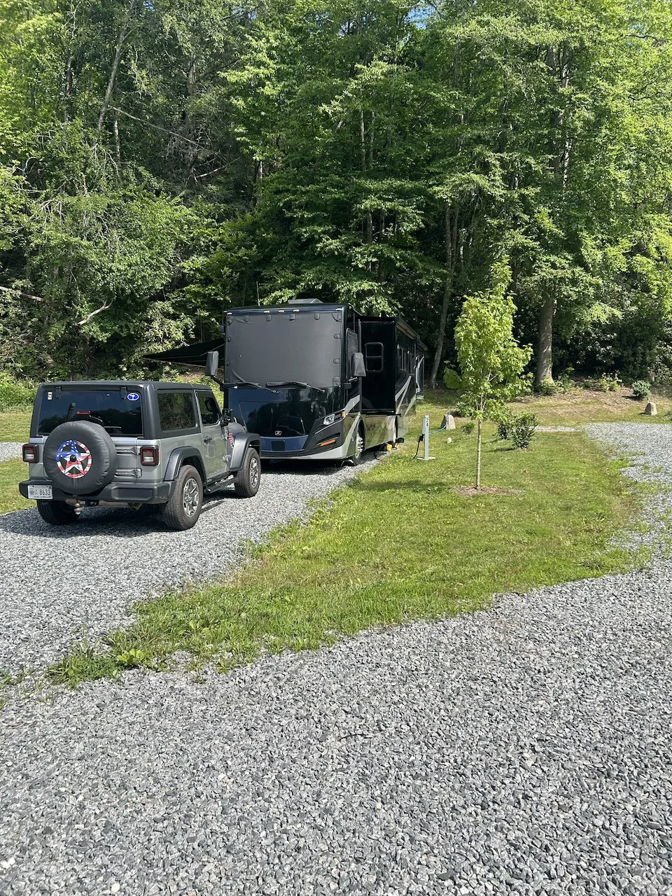 A gray Jeep with a spare tire at the back and a black RV parked on a gravel area surrounded by green grass and trees.