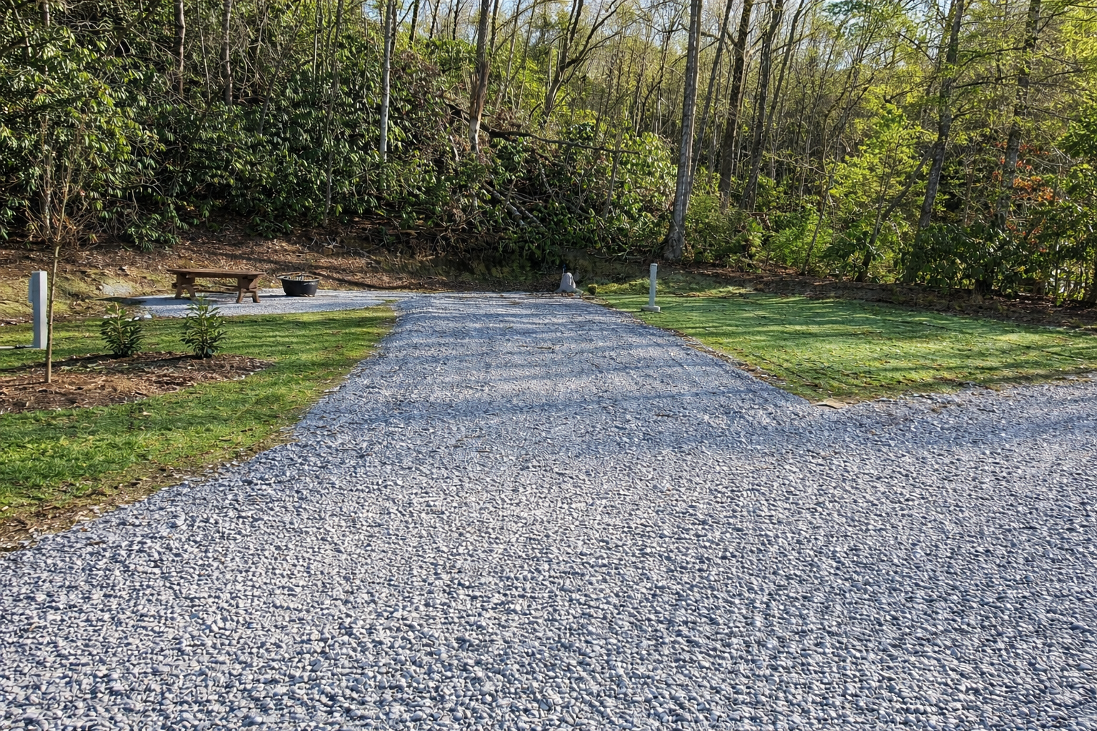 Gravel pathway leading to a meditative area with a bench and a fire pit, surrounded by greenery and trees.
