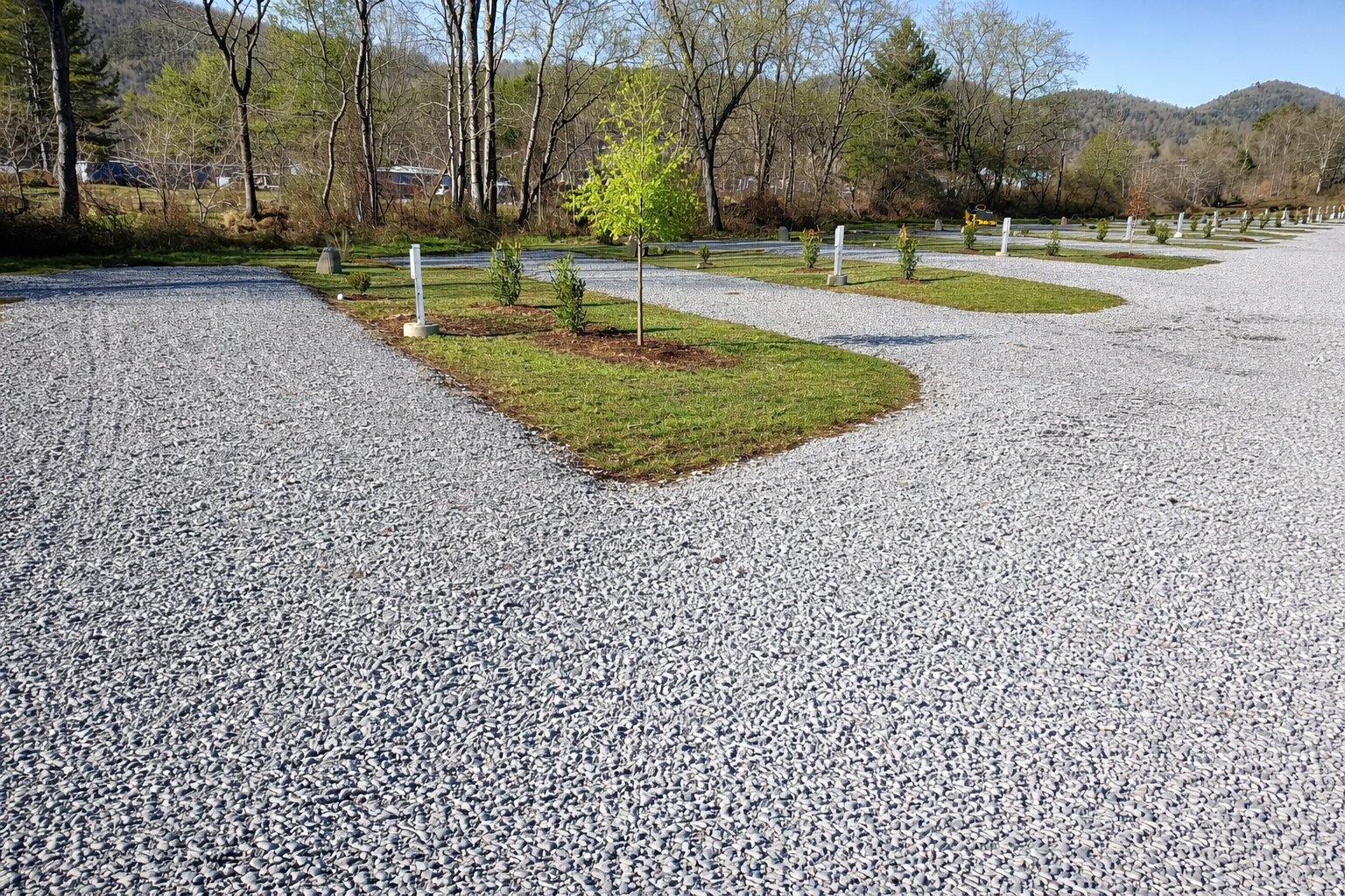 Gravel parking lot with small landscaped islands having young trees and shrubs, surrounded by grass, in a rural, hilly area during daytime.