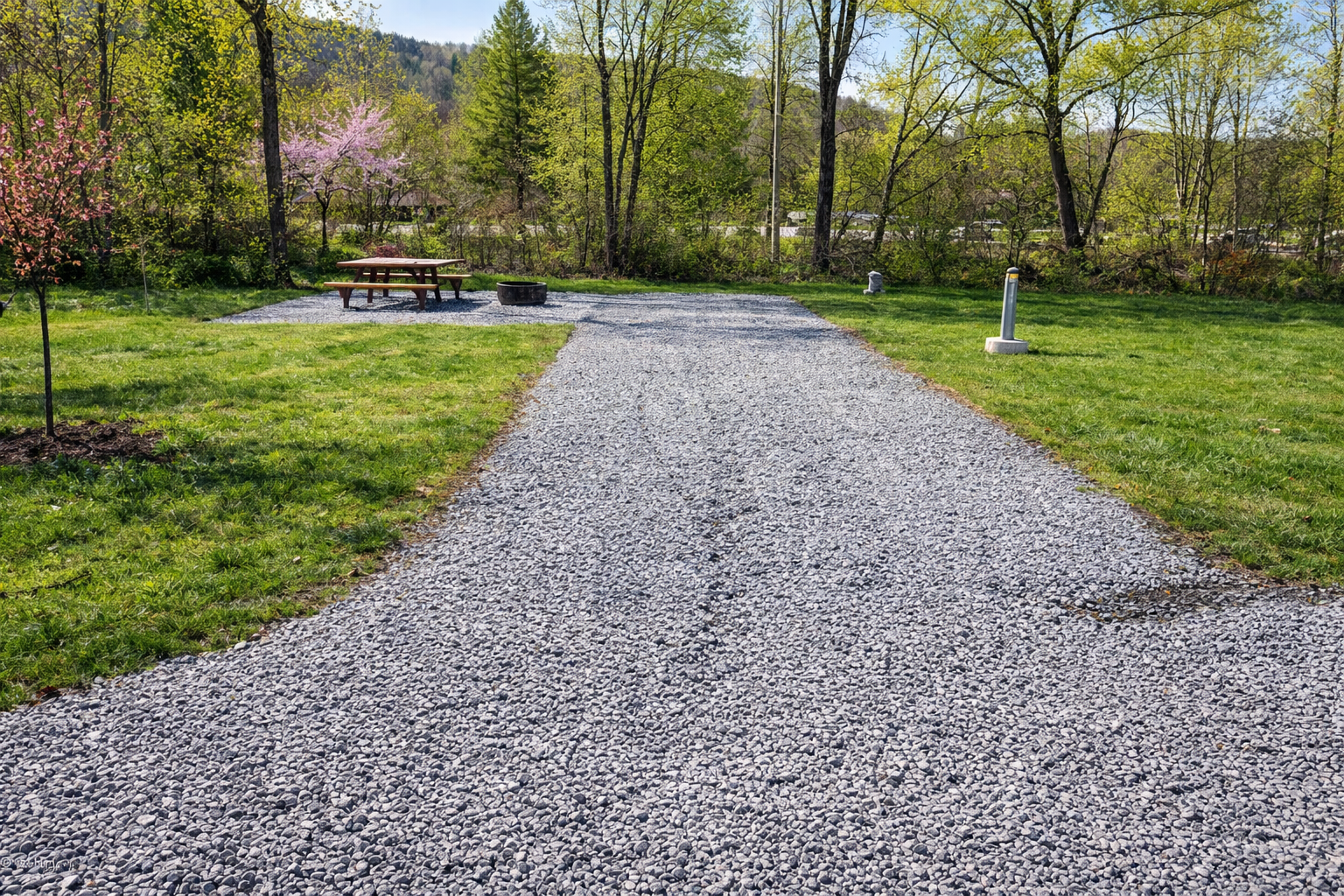 A gravel walking path in a park with green grass on both sides and young trees, some with pink blossoms, under a clear blue sky, with a picnic table and a fire pit in the background.