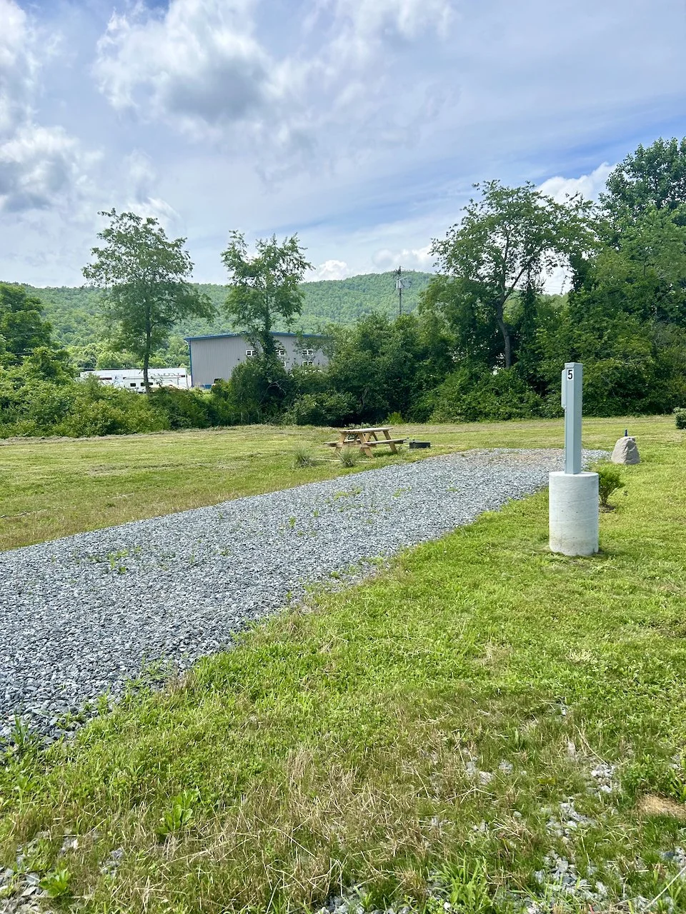 A gravel pathway in a grassy field with a picnic table, trees, and mountains in the background under a partly cloudy sky.