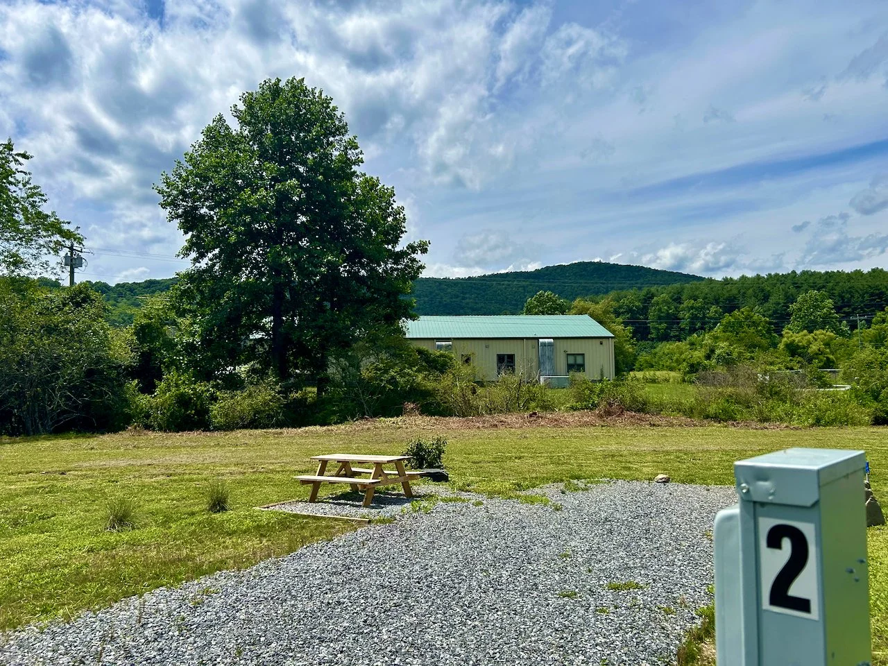 A grassy outdoor area with a gravel path, a small picnic table on a concrete pad, a tree, a metal building with a green roof, and a background of rolling hills and trees under a partly cloudy sky.