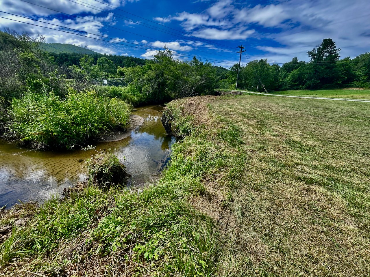 A small stream runs through a grassy area next to a field, with green trees and hills in the background under a partly cloudy blue sky.