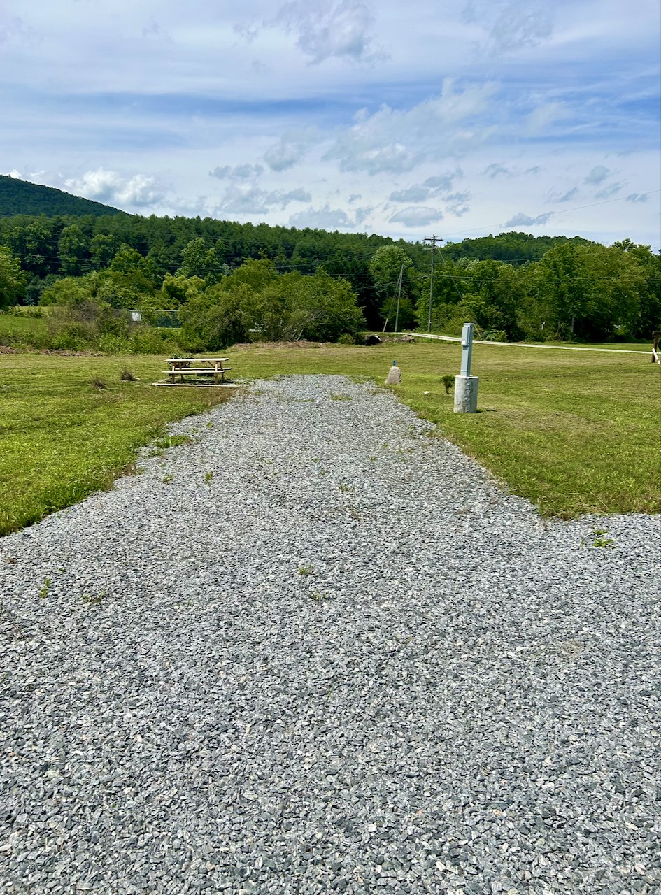 Gravel pathway in a grassy park with a picnic table, trees, hills, and a cloudy sky in the background.