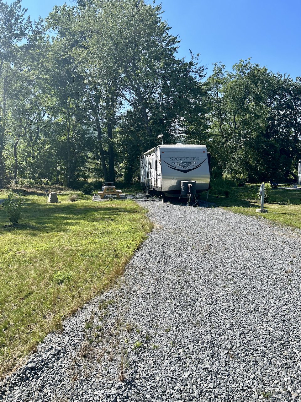 A gravel driveway leading to a gray travel trailer labeled 'Sportsmen' parked in a wooded area with trees and a picnic table nearby.