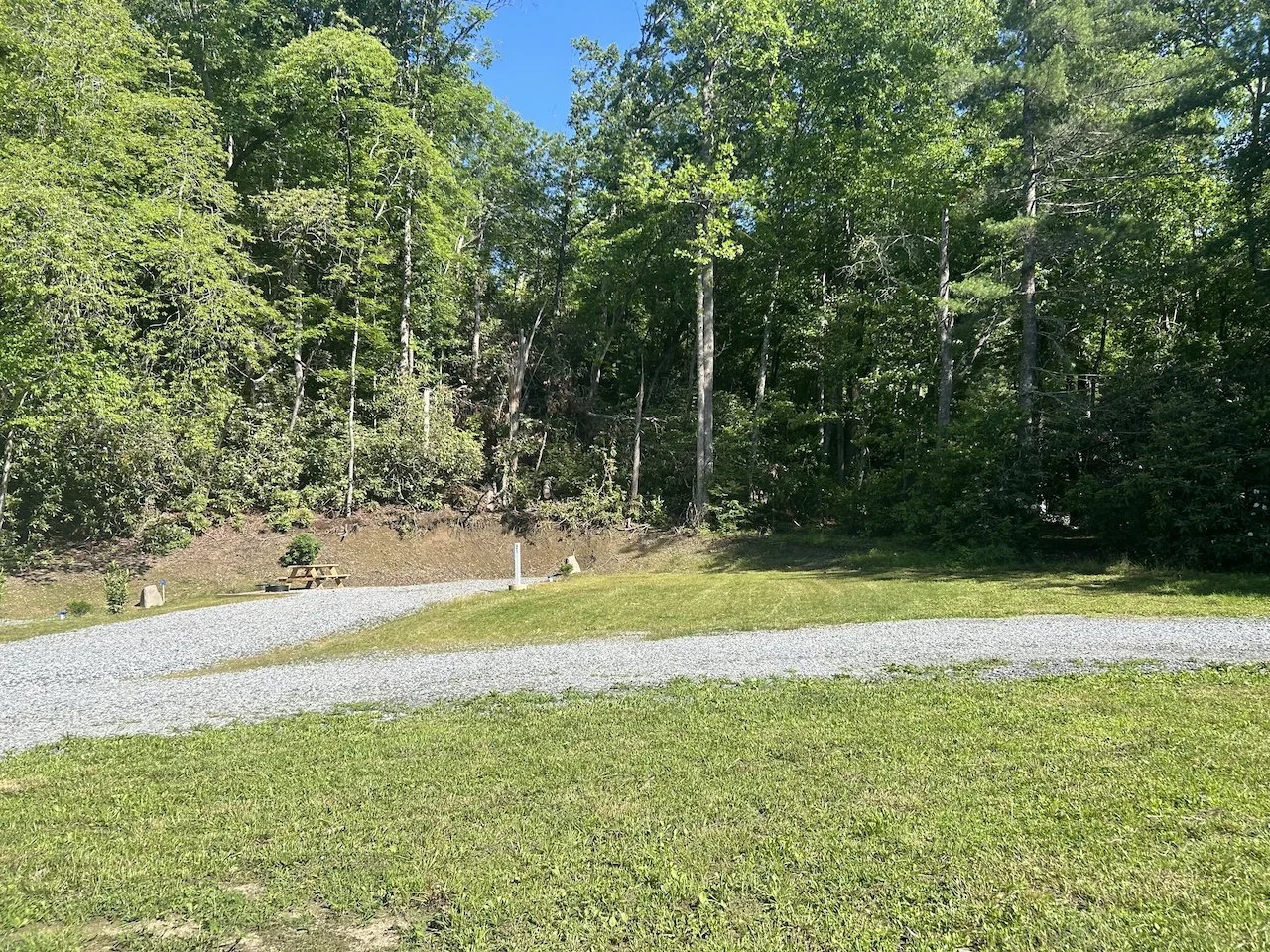 A grassy outdoor area with a gravel pathway and a picnic table near a wooded area under a blue sky.
