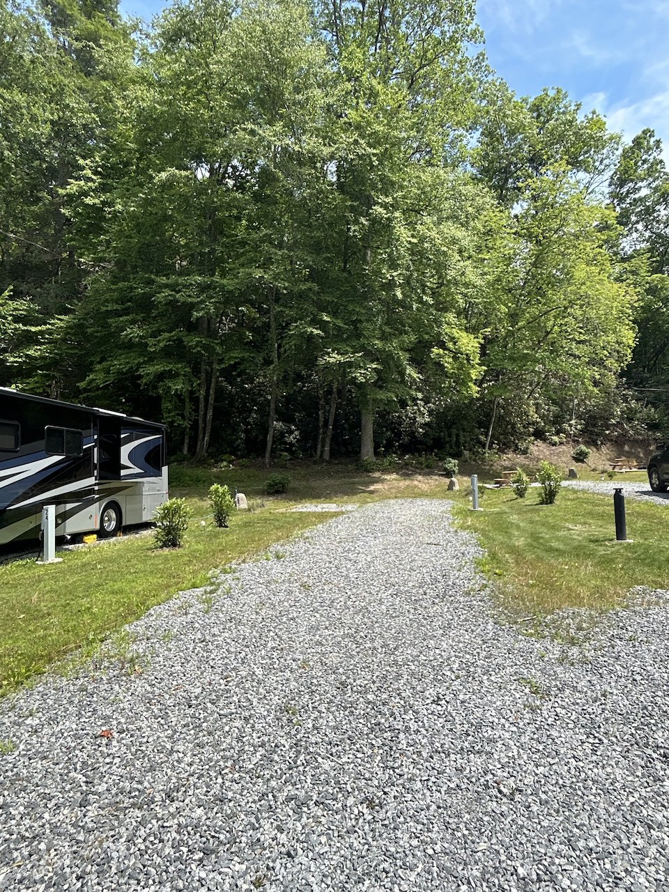 Camping site with gravel path, parked RV, small bushes, and trees in the background on a sunny day.