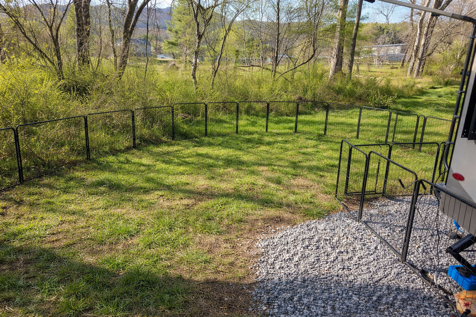 A fenced-in yard with black metal panels, surrounded by trees and greenery, with a gravel pathway on one side, and a trailer partially visible on the right.